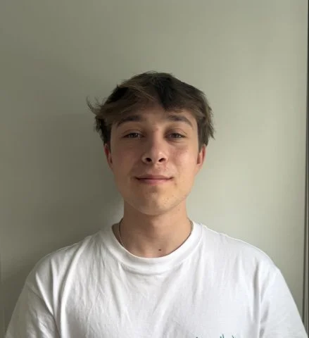 A young man with light brown hair wearing a white t-shirt standing in front of a plain wall.
