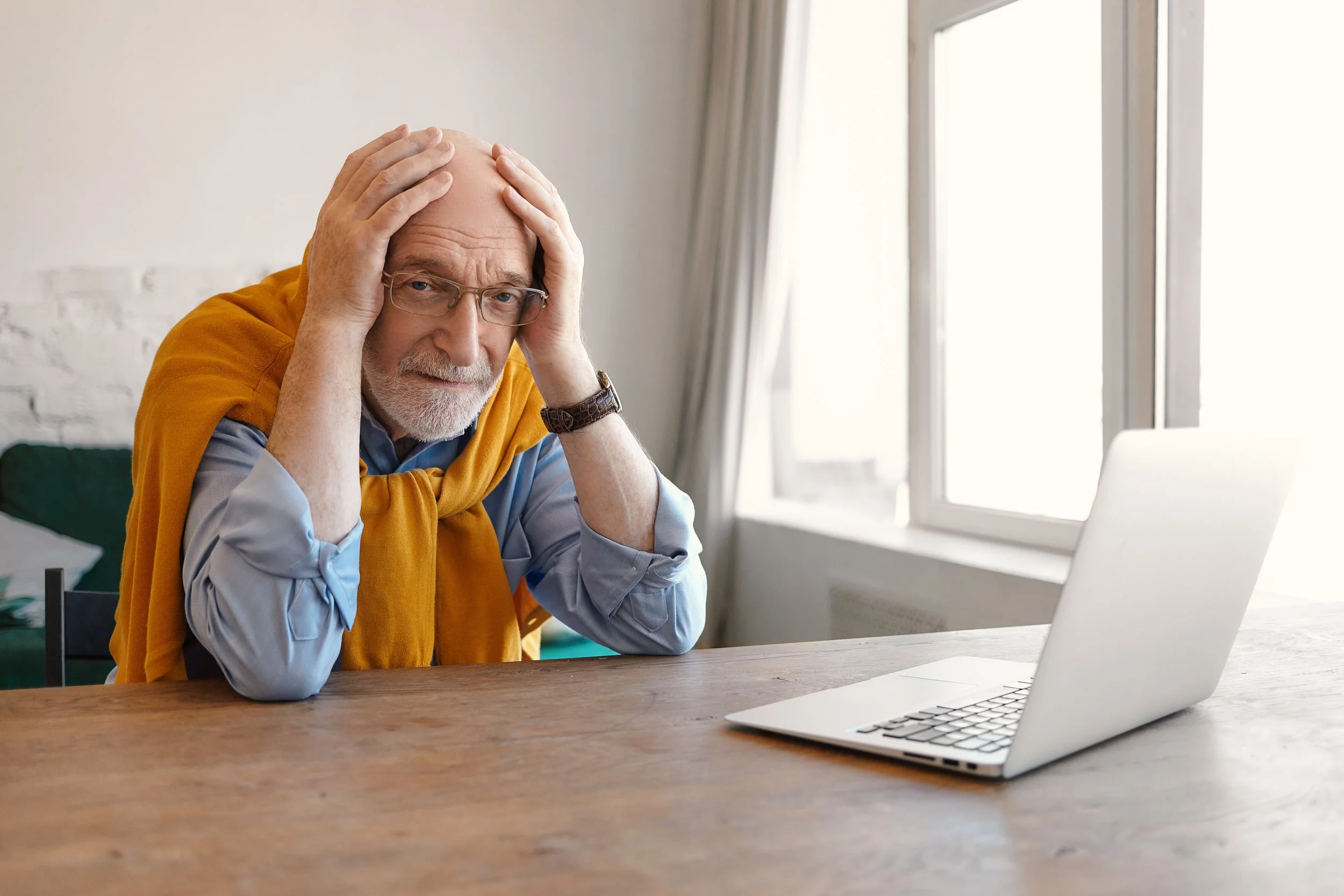 An elderly man with glasses and a gray beard looks distressed, holding his head with both hands at a wooden desk with a laptop, in a bright room with large windows.