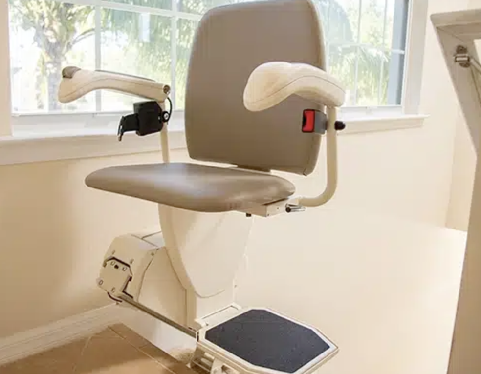 A beige stair lift chair with wooden armrests and a backrest, installed next to a window with greenery outside, in a room with beige walls and tiled floor.