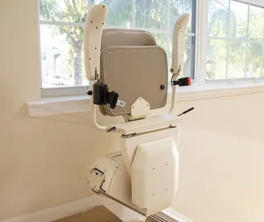 An adjustable medical examination or imaging chair with arm supports, positioned in front of a window in a room with beige walls.