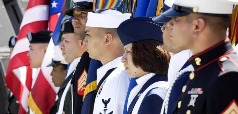Line of military personnel in uniform, standing at attention during a ceremony, with American and other flags behind them.