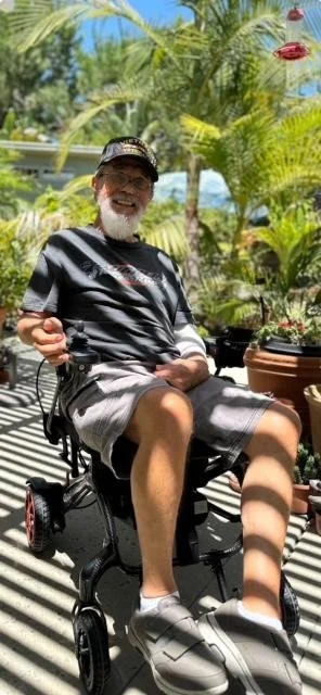 An elderly man with a beard smiling and sitting in a motorized wheelchair outdoors, surrounded by lush green plants and bright pink flowers, with a sunny sky in the background.