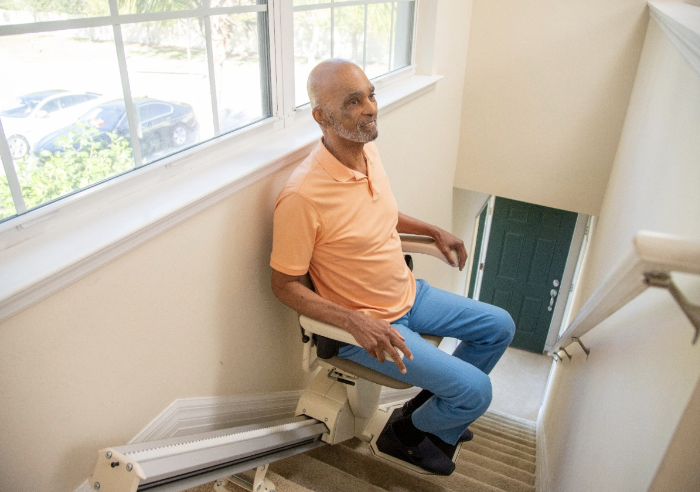 An elderly man with a beard is sitting on a stairlift at the top of a staircase in a home, wearing an orange polo shirt and blue pants, looking out the window with a smile.