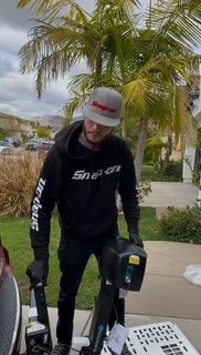 A person in a black hoodie and gray cap standing outdoors, next to a golf cart on a sidewalk with palm trees and houses in the background.