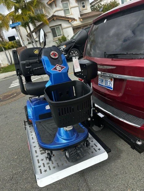Blue mobility scooter with a black basket, parked next to a red vehicle in a parking lot, with residential buildings and trees in the background.