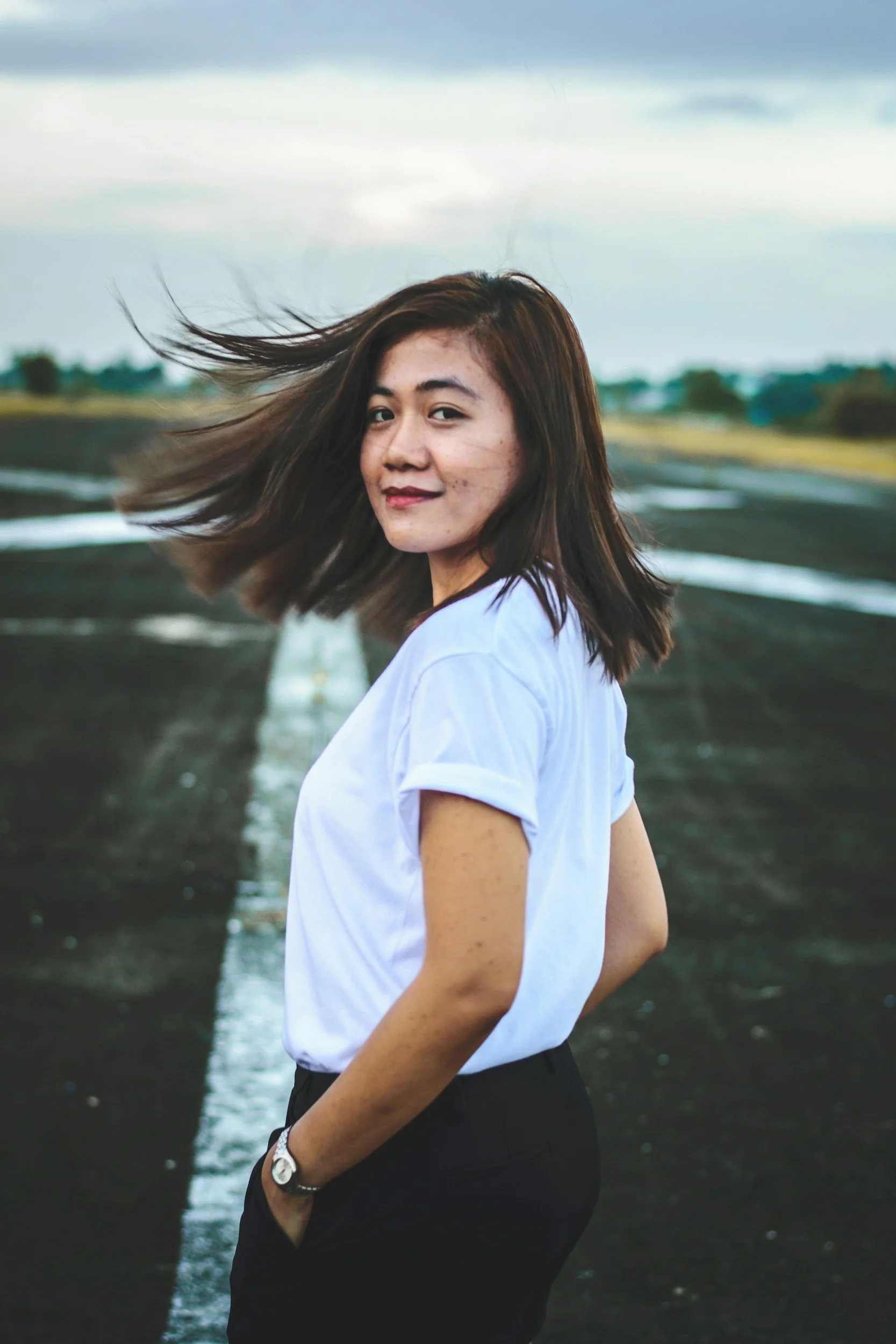 A woman with shoulder-length brown hair wearing a white T-shirt and black pants, standing on an empty road with her hands in her pockets, looking back at the camera with wind blowing her hair.