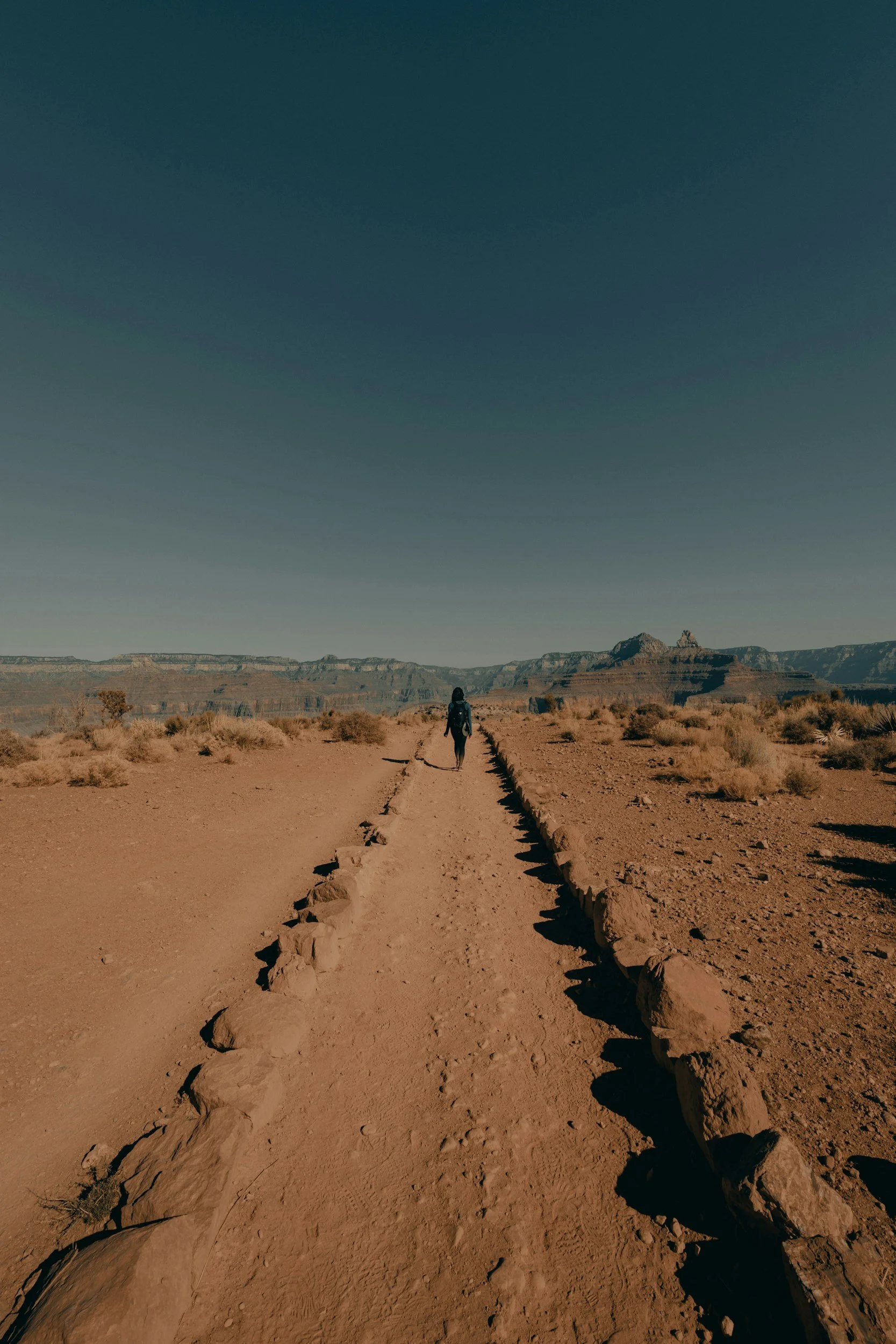 A person walking alone on a dirt desert trail with rocky borders, in a vast arid landscape with distant cliffs and sparse vegetation under a clear blue sky.