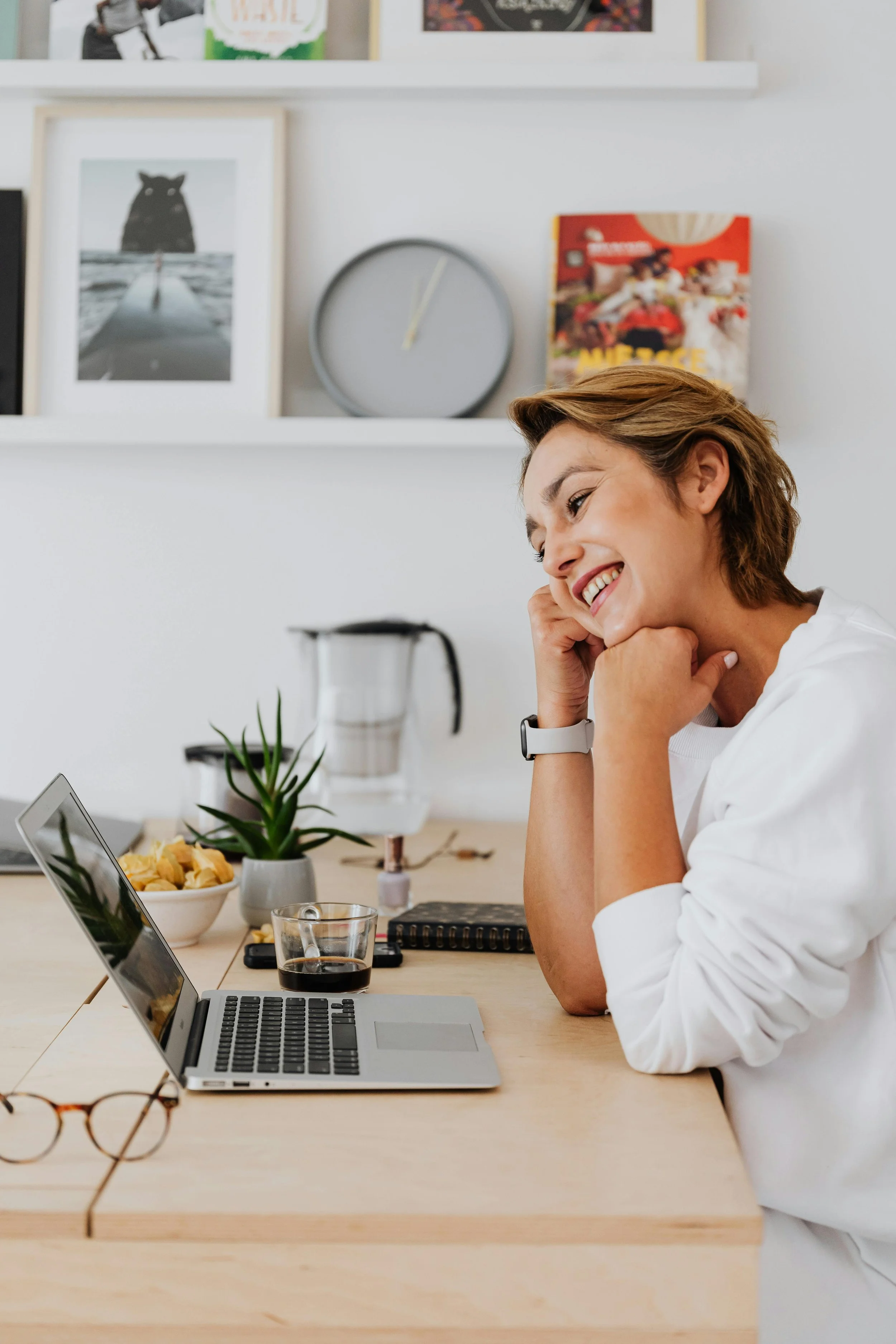 A woman smiling at her laptop at a kitchen table with a bowl of snacks, a glass of soda, and a potted plant.