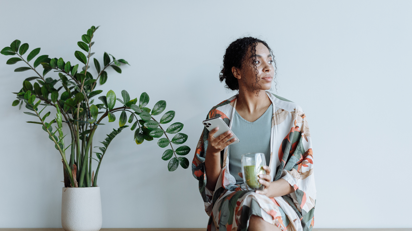 A woman sitting at a table holding her phone and looking into the distance, reflecting overthinking and emotional distraction.