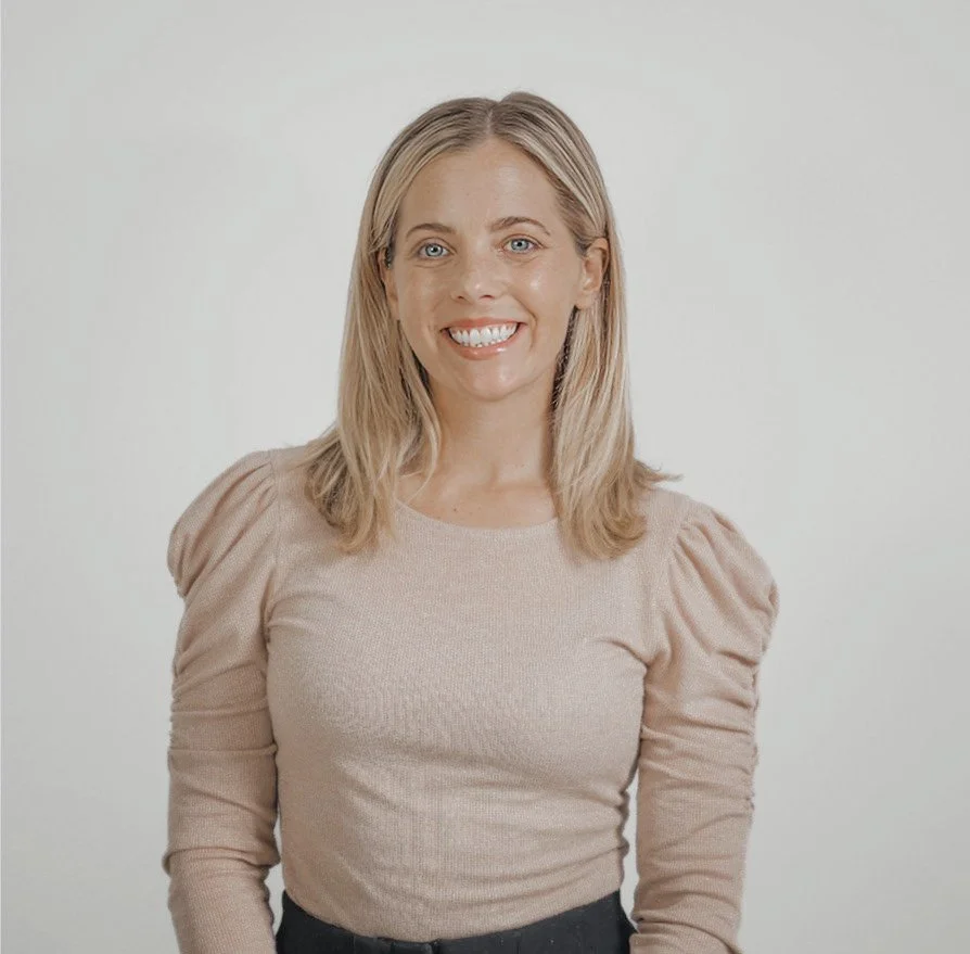 A woman with shoulder-length blonde hair, blue eyes, and a bright smile, wearing a beige long-sleeve top with puffed shoulders, standing against a plain light gray background.