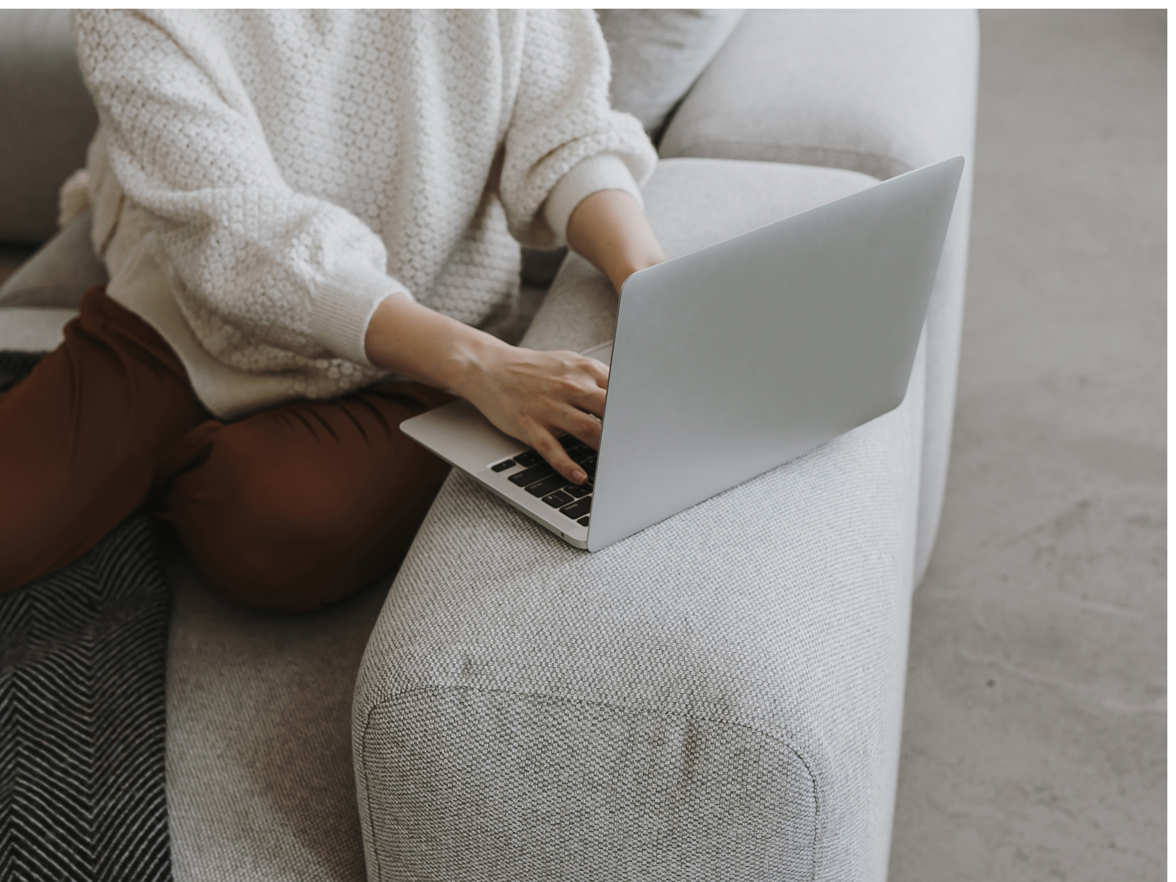 Person using a laptop on a light-colored sofa wearing a white knit sweater and brown pants.