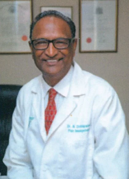 A smiling man in a white medical coat with a red patterned tie, wearing glasses, sitting in an office or clinic.