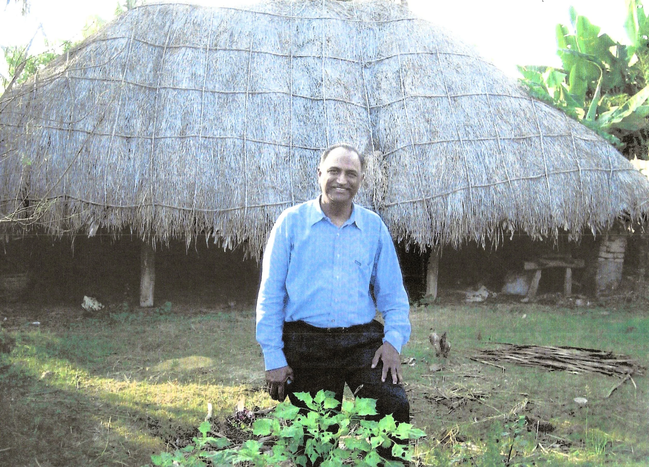 A man in a blue shirt and black pants kneeling on the grass in front of a traditional thatched hut with a curved roof. There are plants in the foreground and trees in the background.