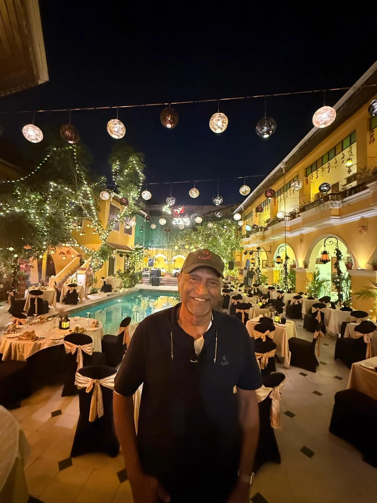 Smiling man in black shirt and cap standing in a festive outdoor restaurant courtyard with tables decorated with black and white chair covers, string lights, and a swimming pool, with a stage and digital sign displaying '2023' in the background at ni