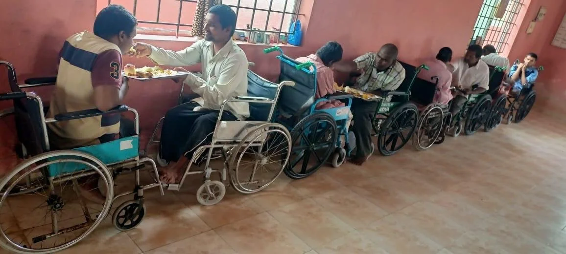 People in wheelchairs receiving food in a dining area of a care facility.