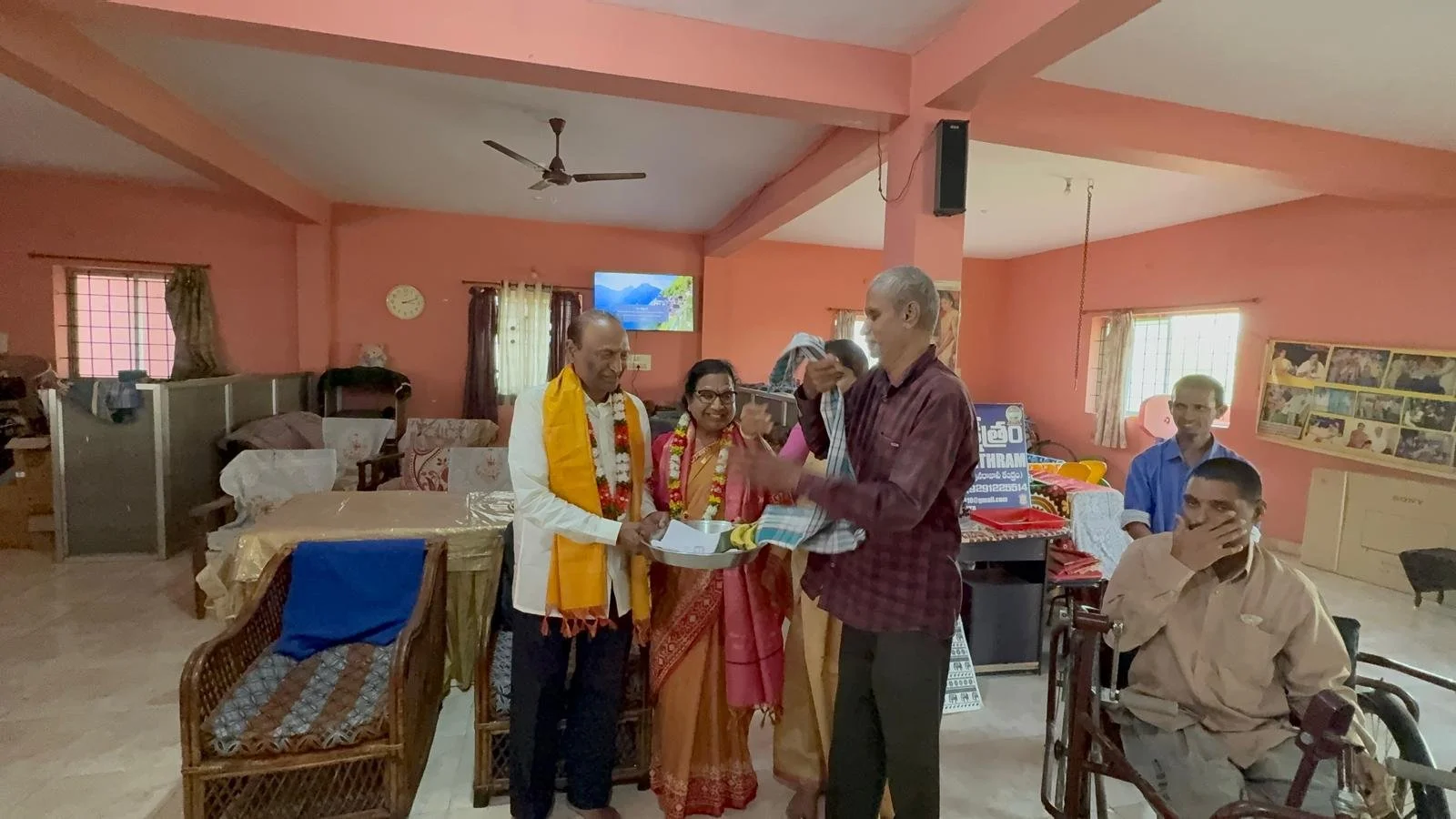 Group of people in a room celebrating or presenting, some wearing traditional Indian attire and floral garlands, with a man in a wheelchair watching, and others behind a table with a tray of items.