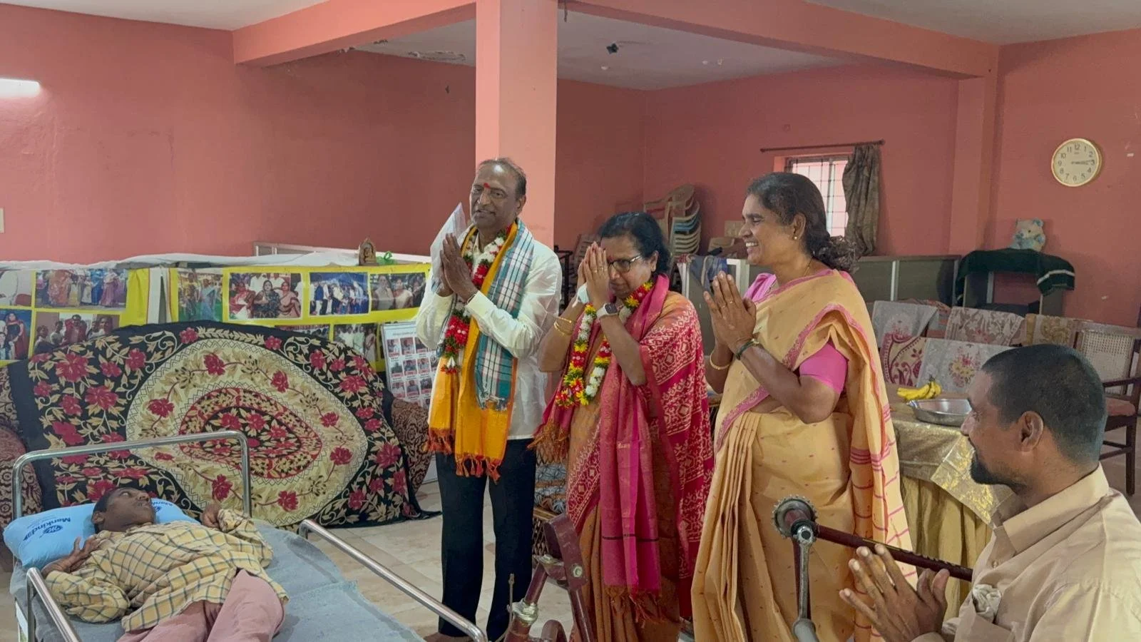 People gathering around a hospital bed with a patient, participating in a religious or spiritual ceremony, with some individuals praying and wearing traditional Indian attire, in a room decorated with photos and banners.
