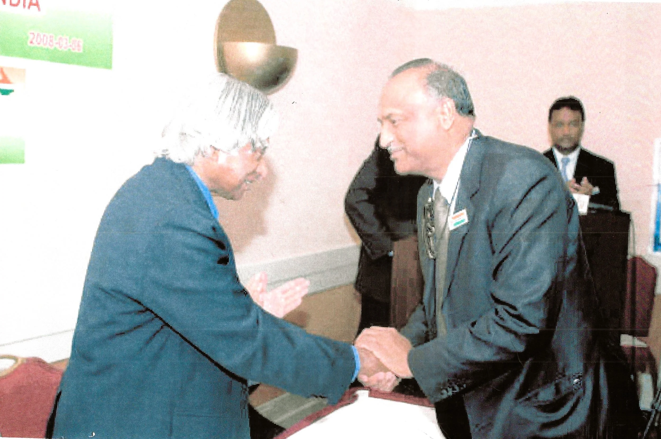 Two men in suits shaking hands in a professional setting, with a third man in the background using a laptop.