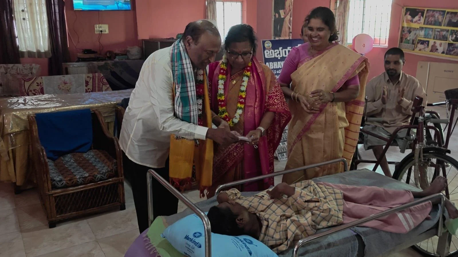 A group of five people, including a man and three women, are gathered around a person lying in a hospital bed. The person in bed is wearing a checked shirt and pink pants. The group appears to be looking at something the man is holding, possibly a ph