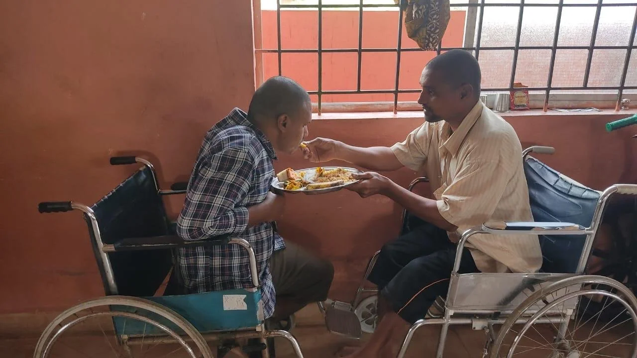 A man in a wheelchair giving food to a young boy in a wheelchair inside a room with a barred window.