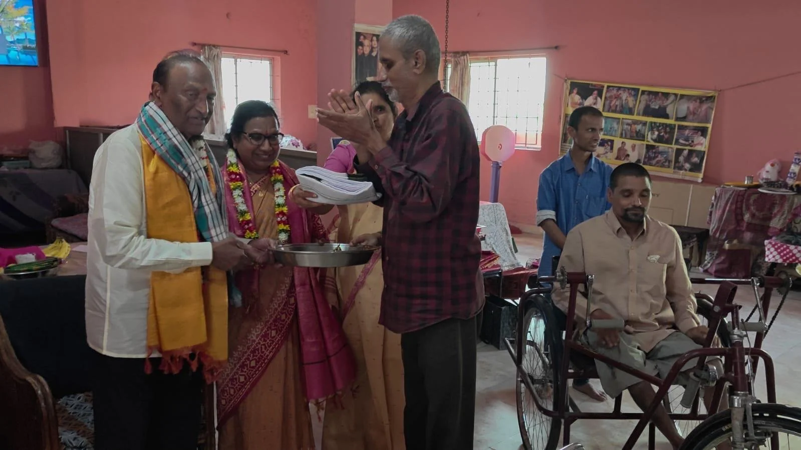Group of people inside a room during a ceremony, with one man in a wheelchair, another man praying with hands together, and women offering a gift or donation.