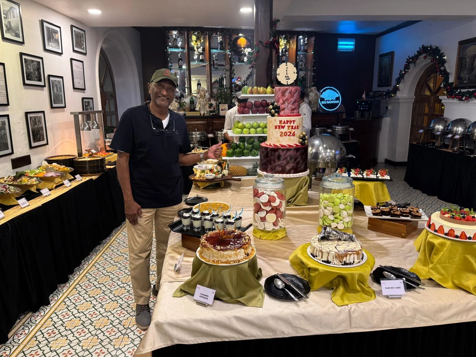 A man standing next to a table with various cakes and desserts at a celebration in a restaurant or hotel banquet hall, decorated for New Year 2026
