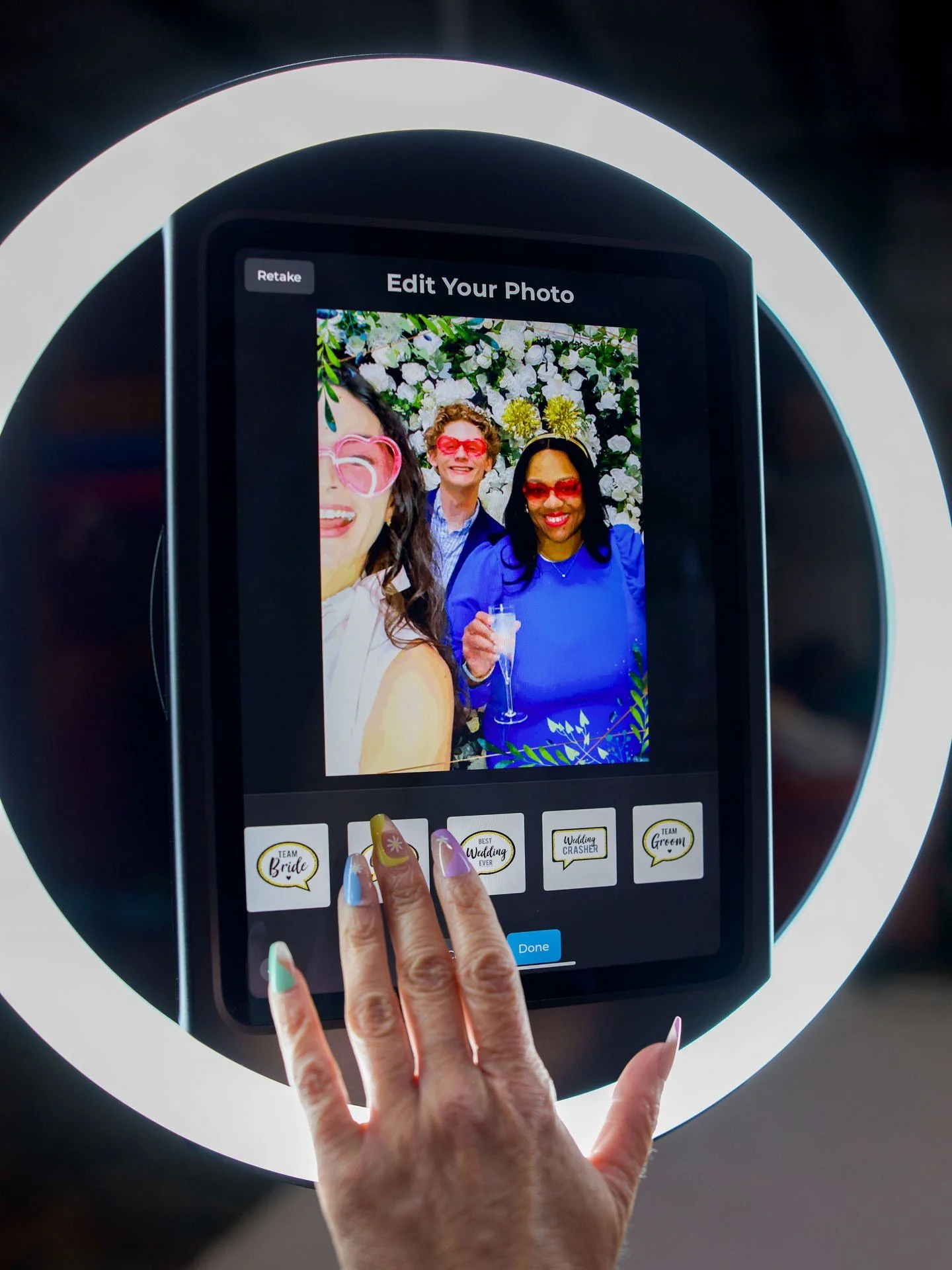 A person's hand with colorful, decorated nails interacts with a photo booth touchscreen, which shows a smiling group of three diverse women posing with sunglasses and drinks, with options to add playful photo stickers.
