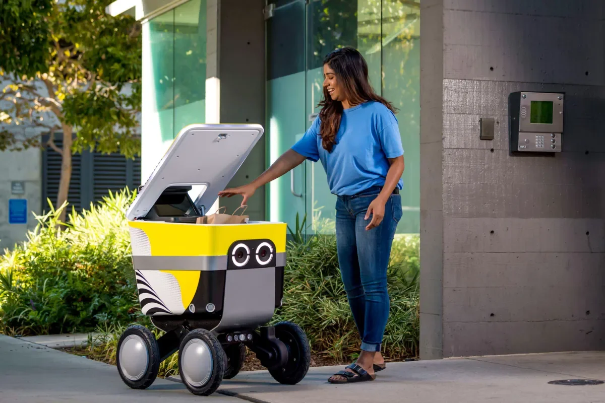 A woman in a blue shirt and jeans interacts with a small, robot delivery vehicle that has a box on top. The vehicle has large wheels, a black and yellow design with cartoonish eyes, and the woman is standing outdoors near a modern building with greenery in the background.