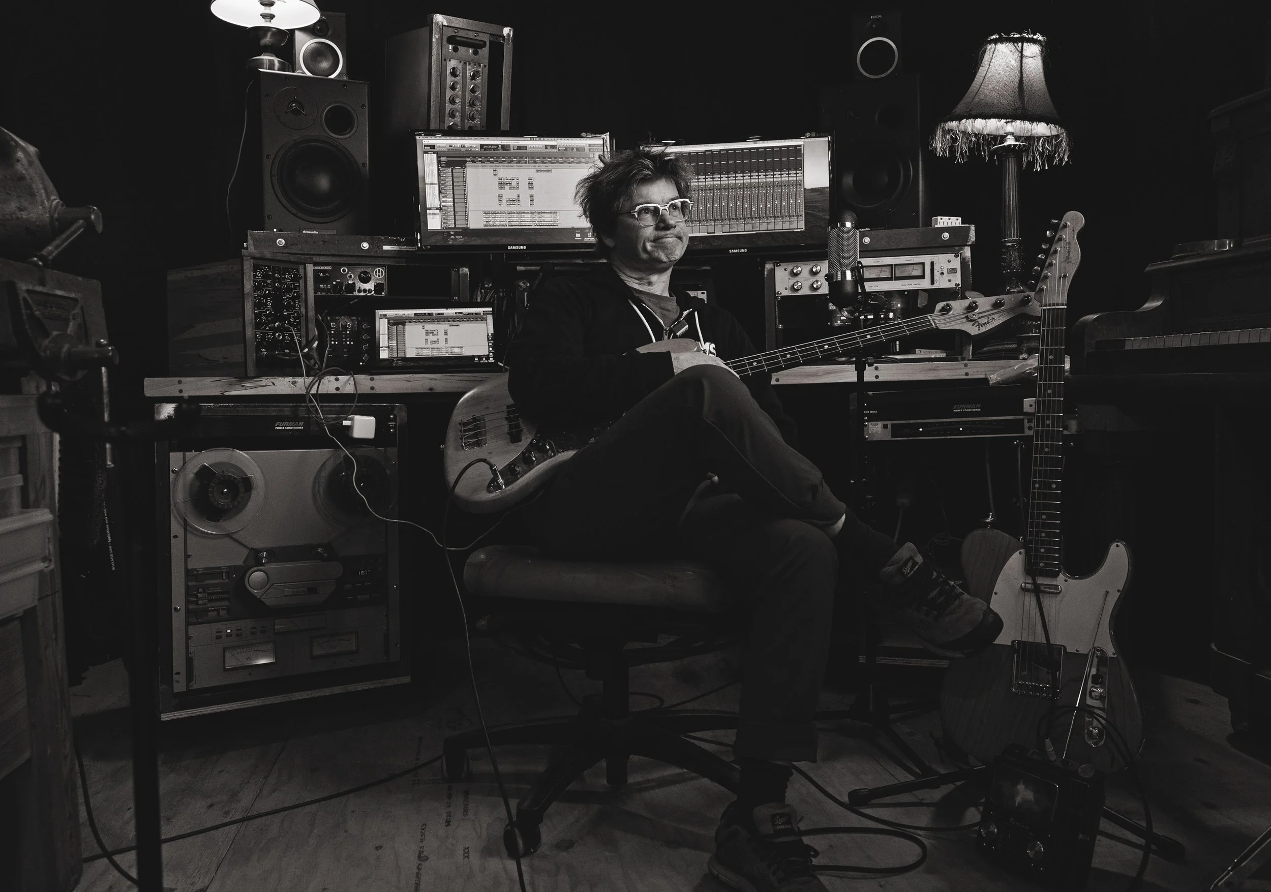 Black and white photo of a man sitting in a recording studio surrounded by audio equipment, two monitors, and musical instruments including an electric guitar and a keyboard.