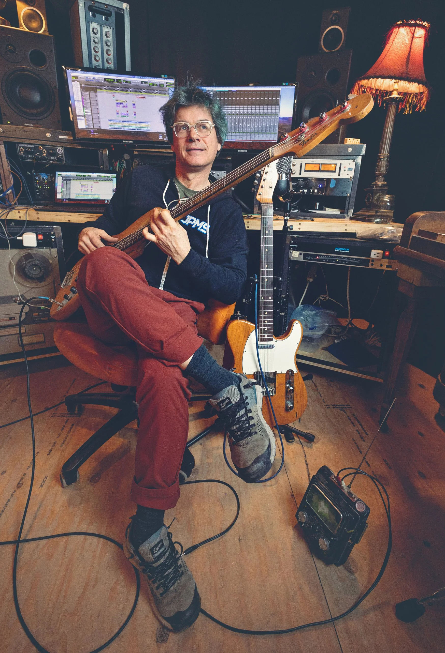 A man with glasses sits in a recording studio holding a bass guitar. There are two other guitars behind him, one upright in a stand. The studio is filled with sound equipment and monitors, with a warm lamp on the right side.