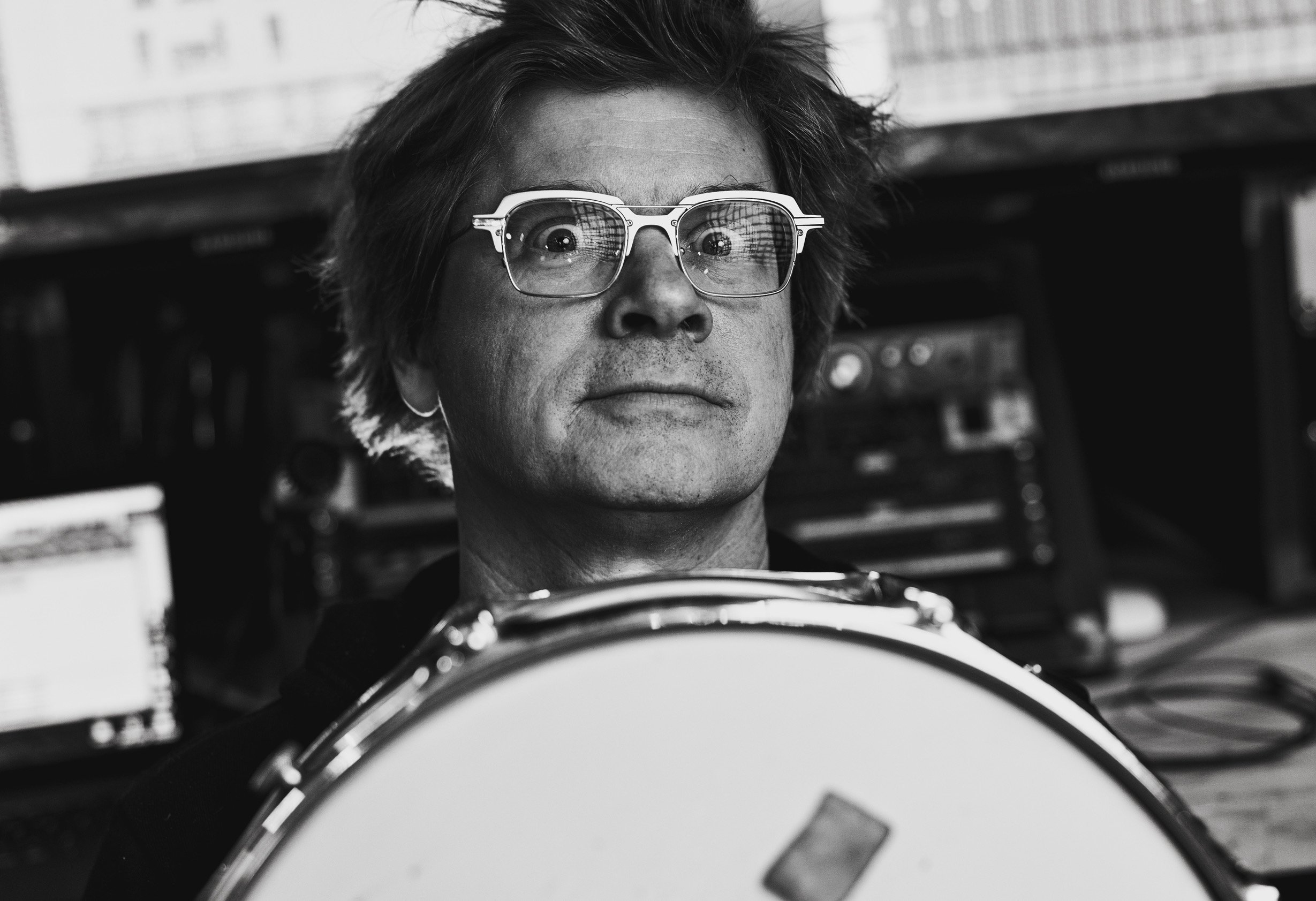 Black and white photo of a man with glasses holding a drum in a music studio.