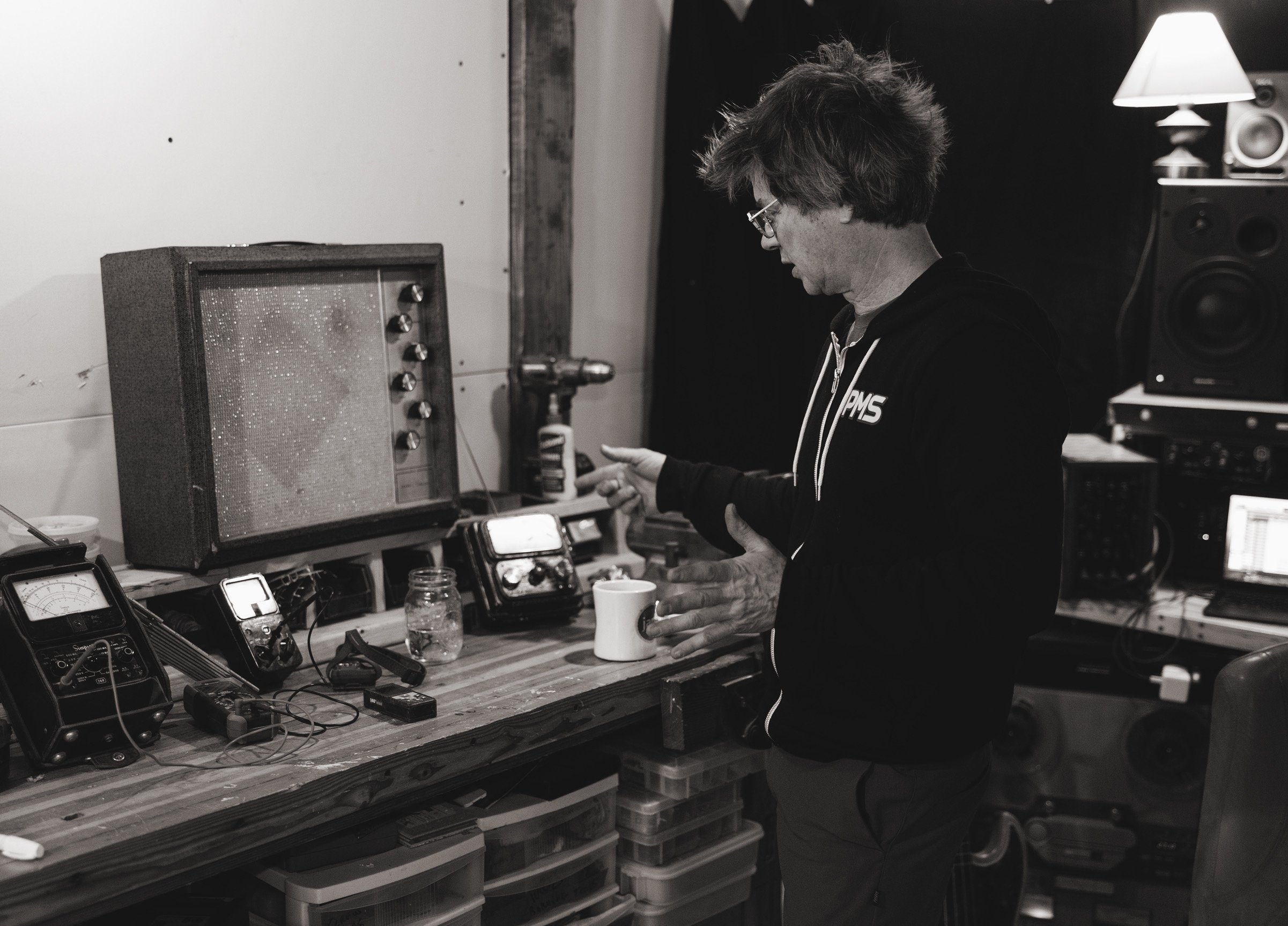 A man with glasses and messy hair standing in a music studio workspace, looking at electronic equipment and a mug on a wooden workbench, surrounded by audio gear and monitors.