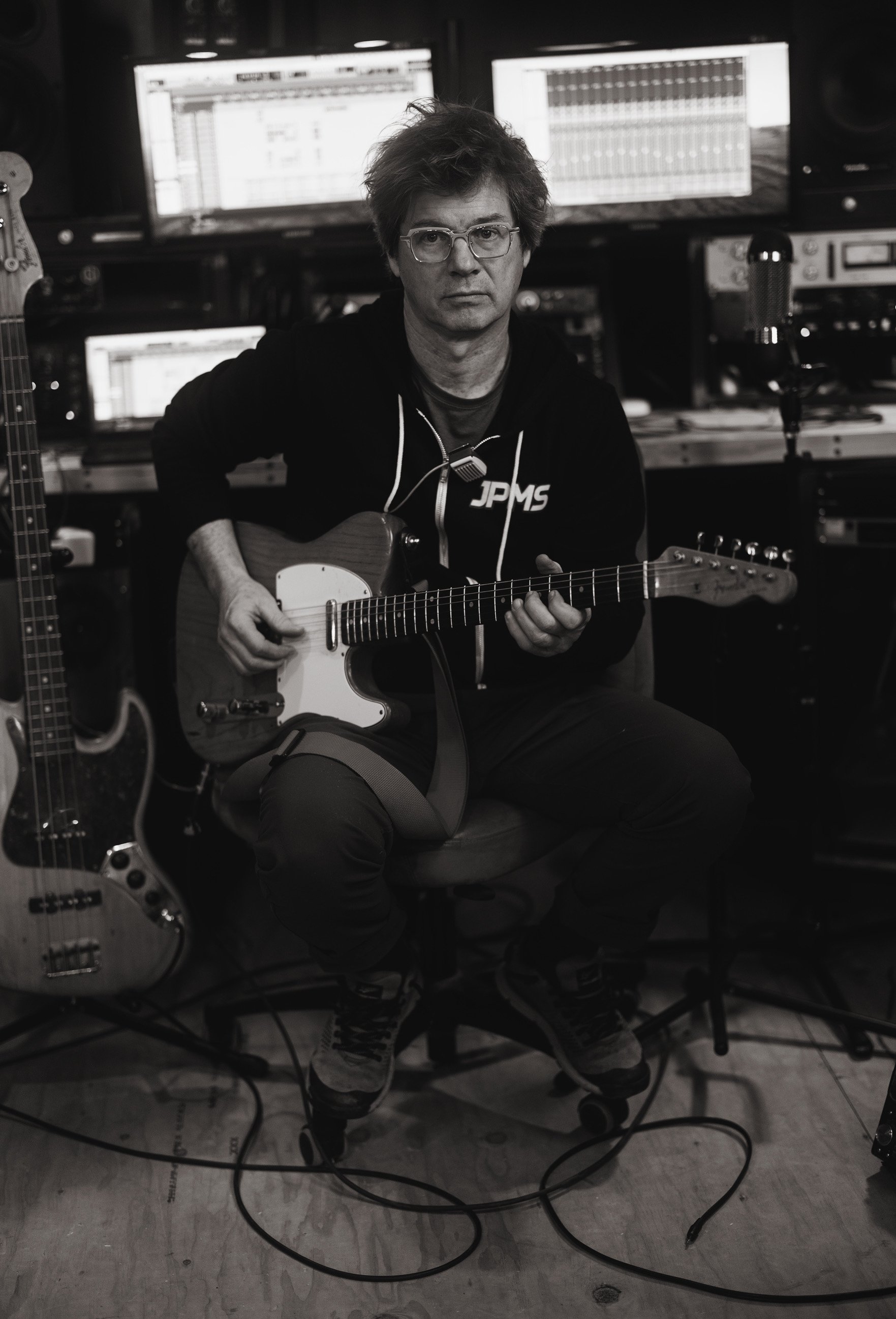 A man with glasses playing an electric guitar in a music studio. The background includes multiple computer screens and music equipment.