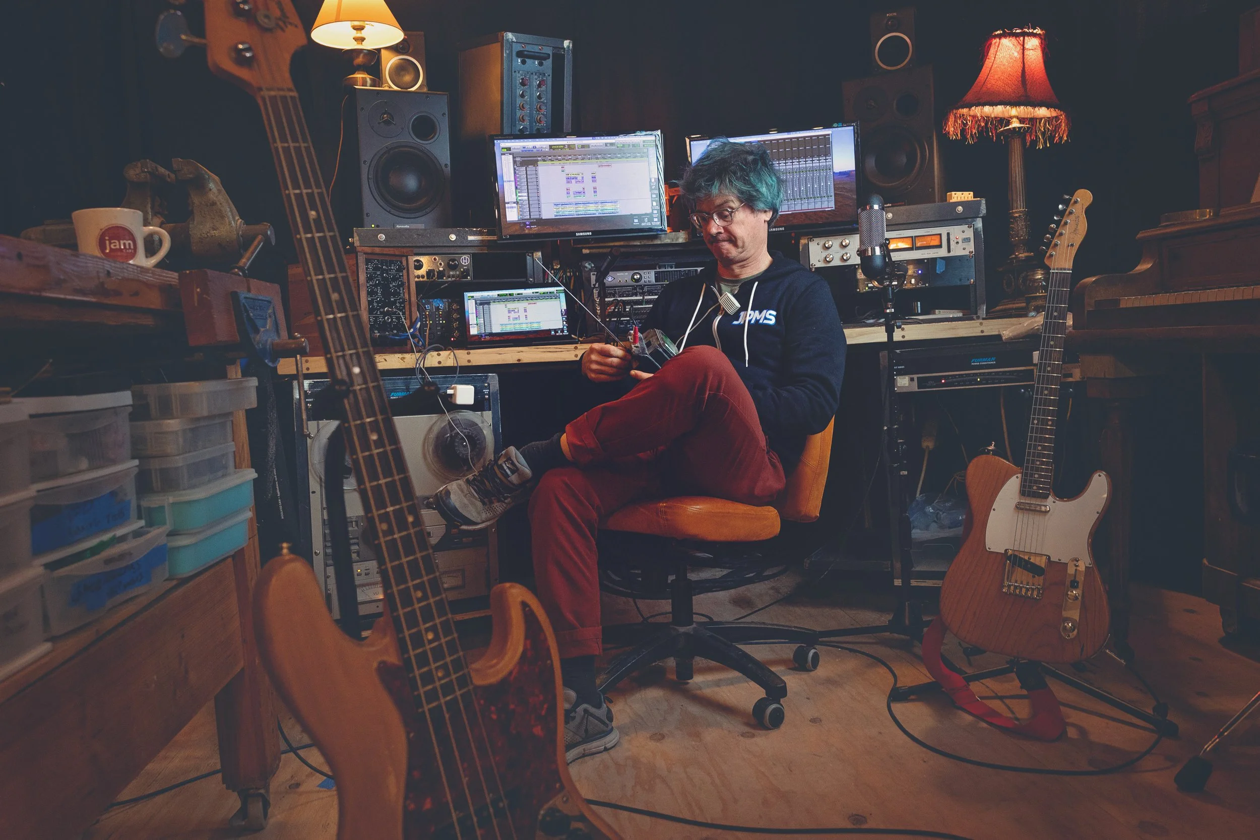 A person with teal hair and glasses is sitting in a music studio surrounded by guitars, sound equipment, and computer monitors.