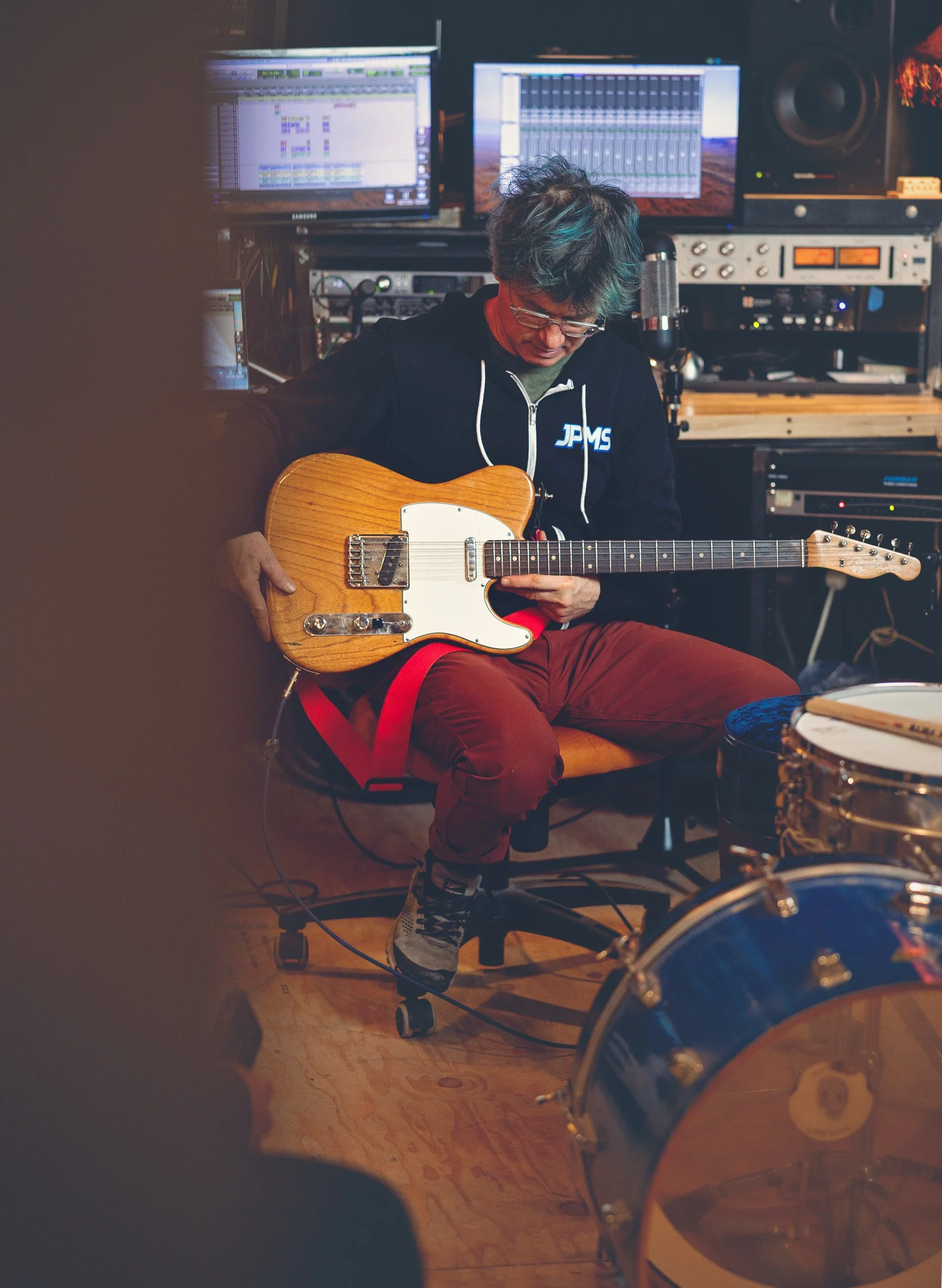 A person with glasses and messy hair sits in a music studio, playing an electric guitar. They wear a black hoodie with "JPMS" on the chest, red pants, and sneakers. The studio has multiple computer monitors, audio equipment, and a drum set in the for