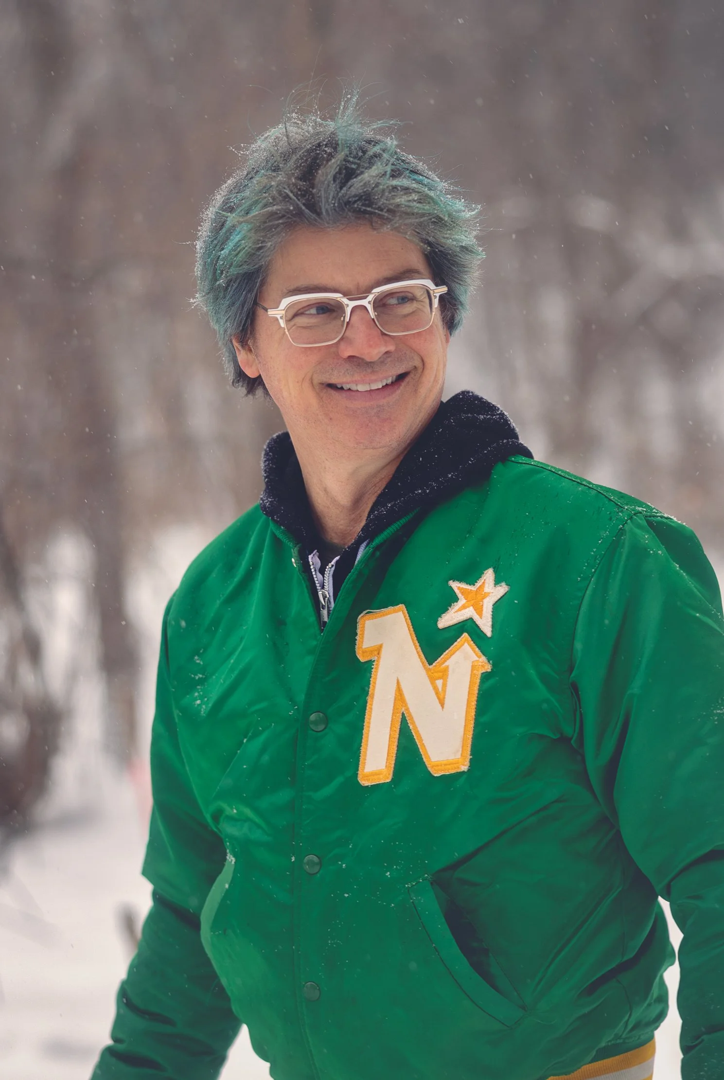 A smiling man with glasses and multicolored hair, wearing a green jacket with an 'Northstars' patch, outdoors in the snow.