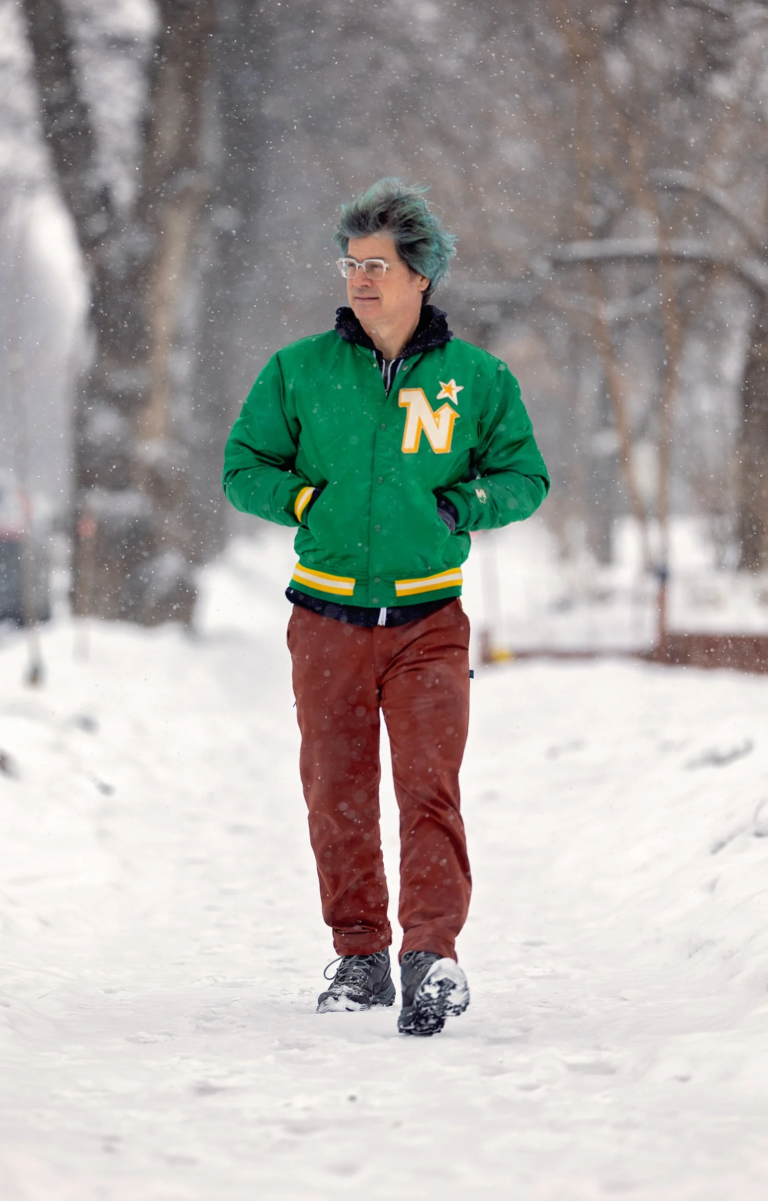 A man walking on snow-covered ground during snowfall, wearing glasses, a green jacket with a large 'Northstars' patch, red pants, and black shoes.