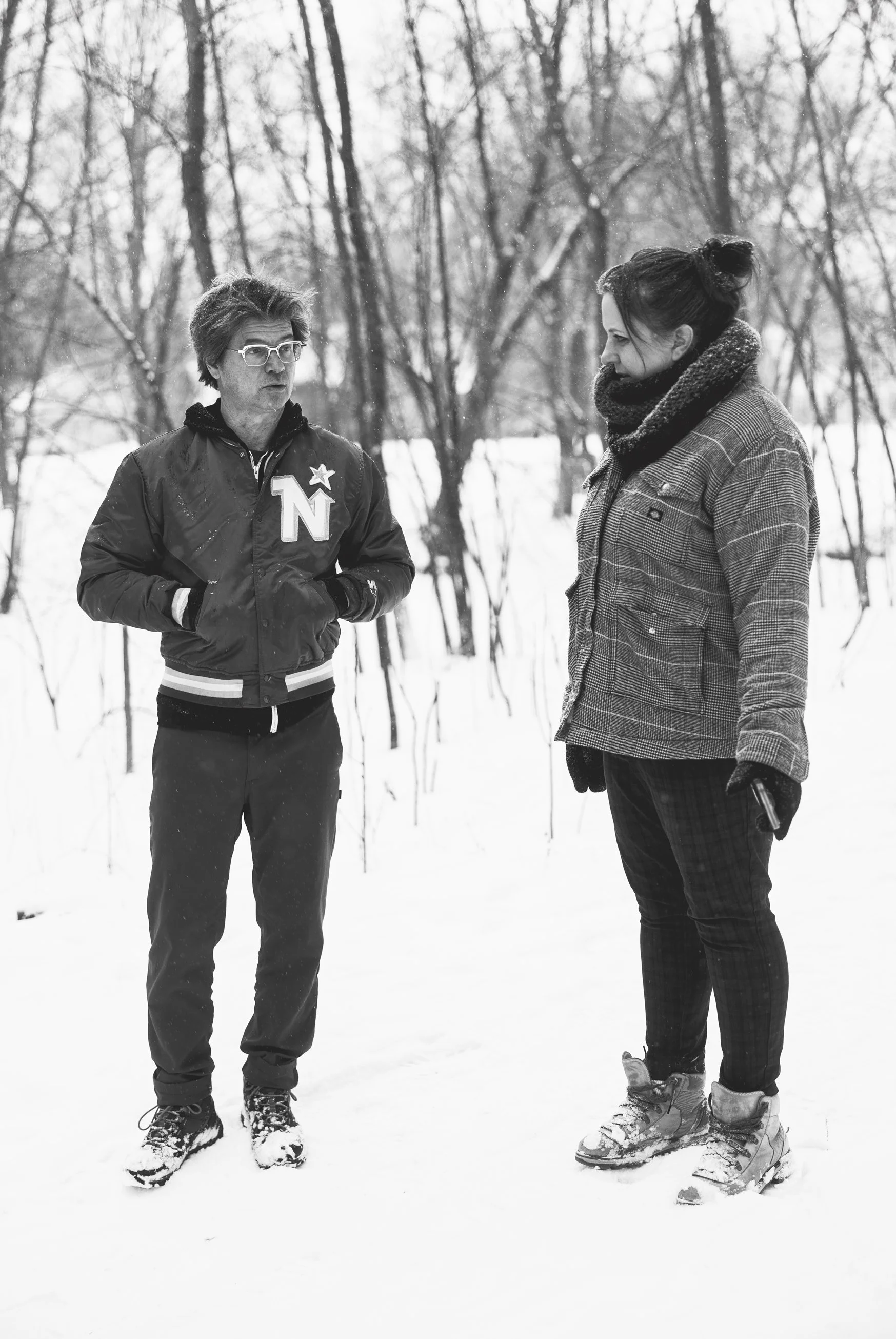 Two people talking outdoors in a snowy forest, one with glasses wearing a jacket with an 'Northstars' patch, the other with a scarf and holding a phone.