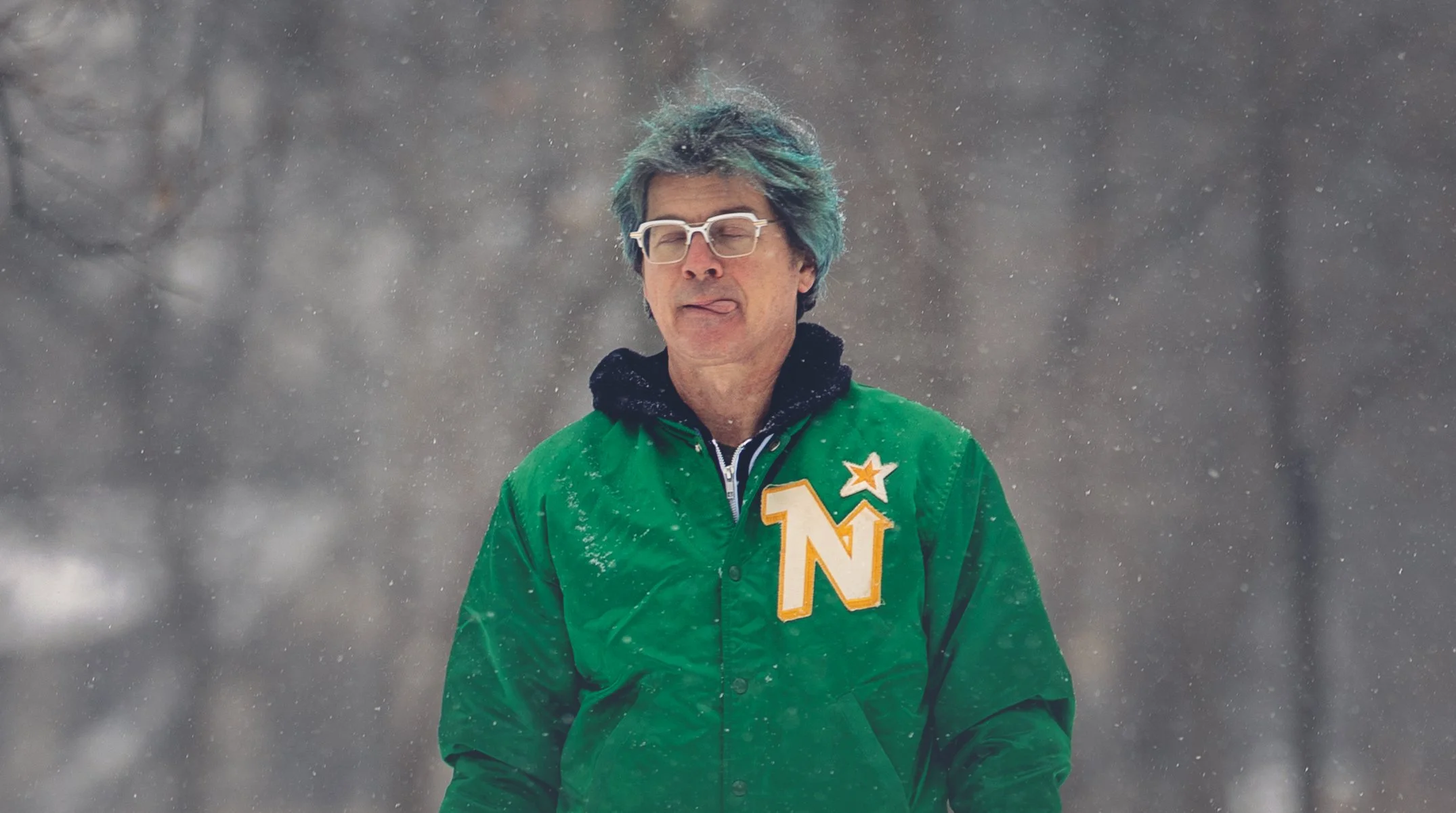 A man with glasses and colorful blue and green hair standing in the snow, wearing a green jacket with a large 'Northstars' patch.
