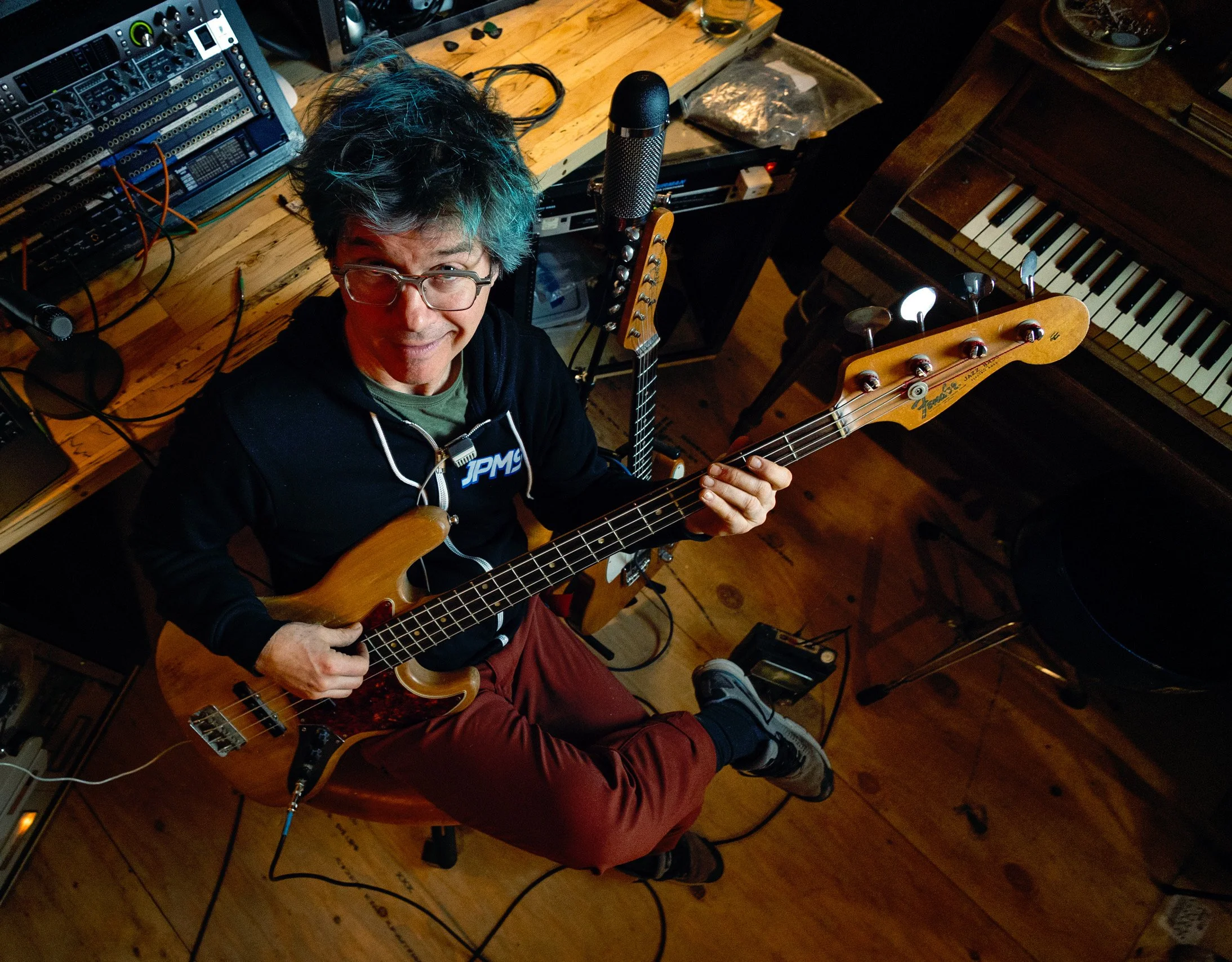 A man with glasses and colorful hair playing a bass guitar in a music studio, surrounded by musical equipment including a keyboard, microphone, and amplifier.