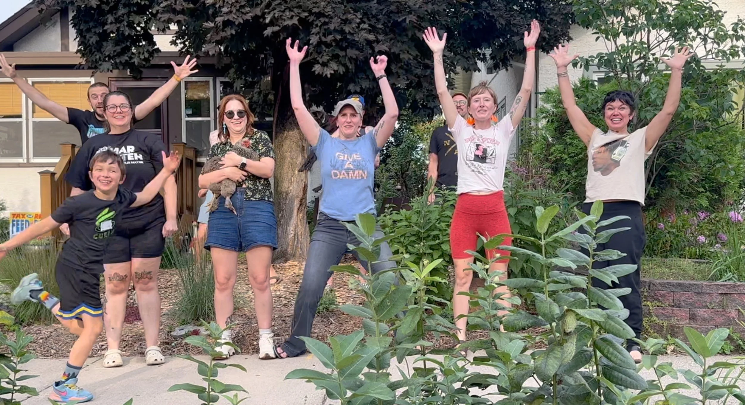 A diverse group of eight people, including children and adults, standing outdoors in front of a house and smiling with arms raised in celebration. One woman is holding a small animal, possibly a kitten.