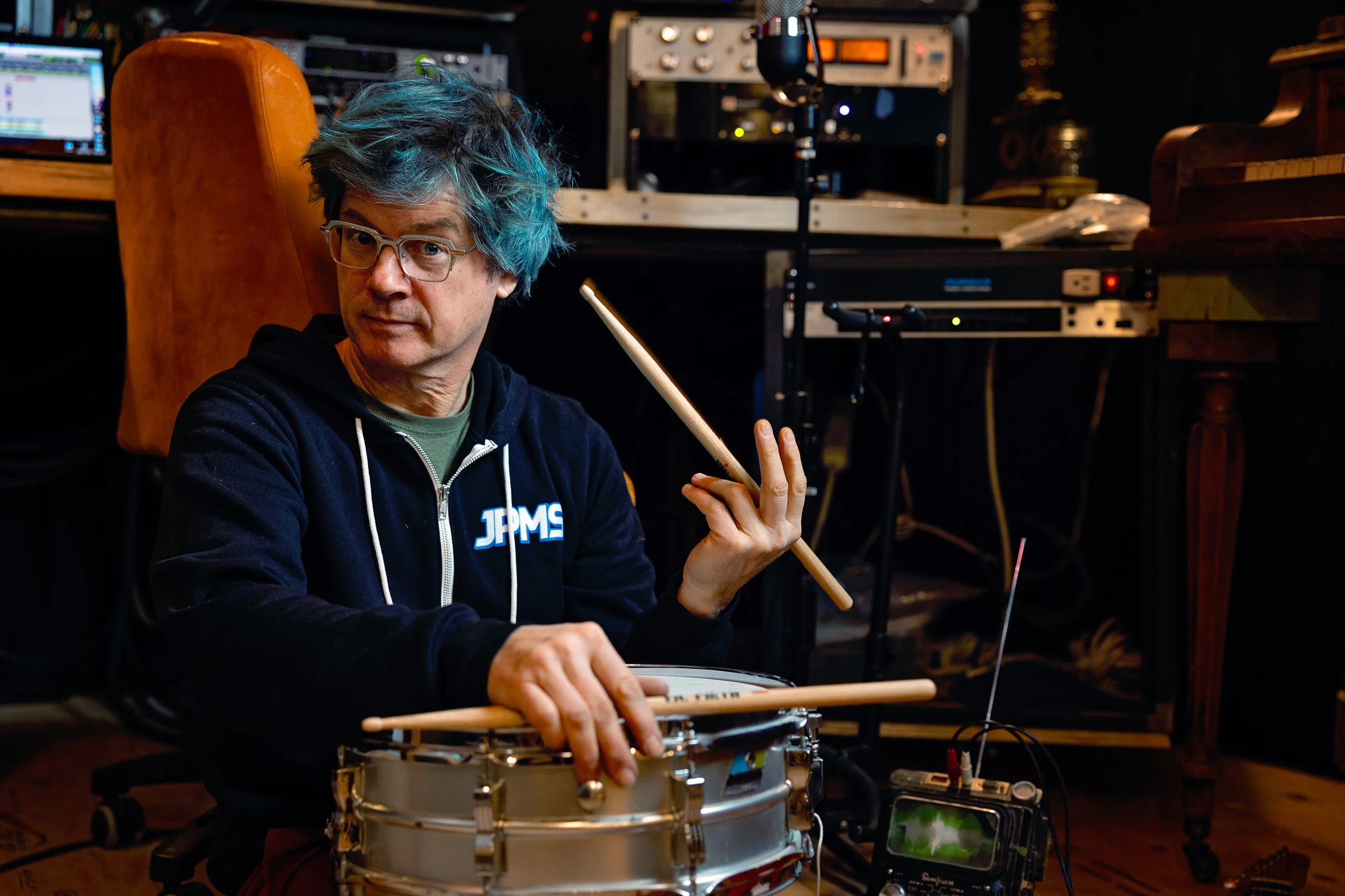 A man with blue hair wearing glasses and a black hoodie sitting in a recording studio with drums, holding drumsticks.