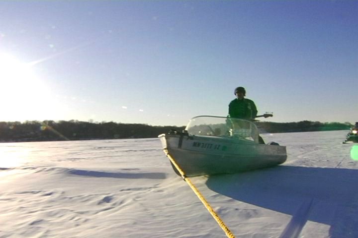 Person on a small boat on a frozen lake, holding a bass guitar during daytime with clear sky.