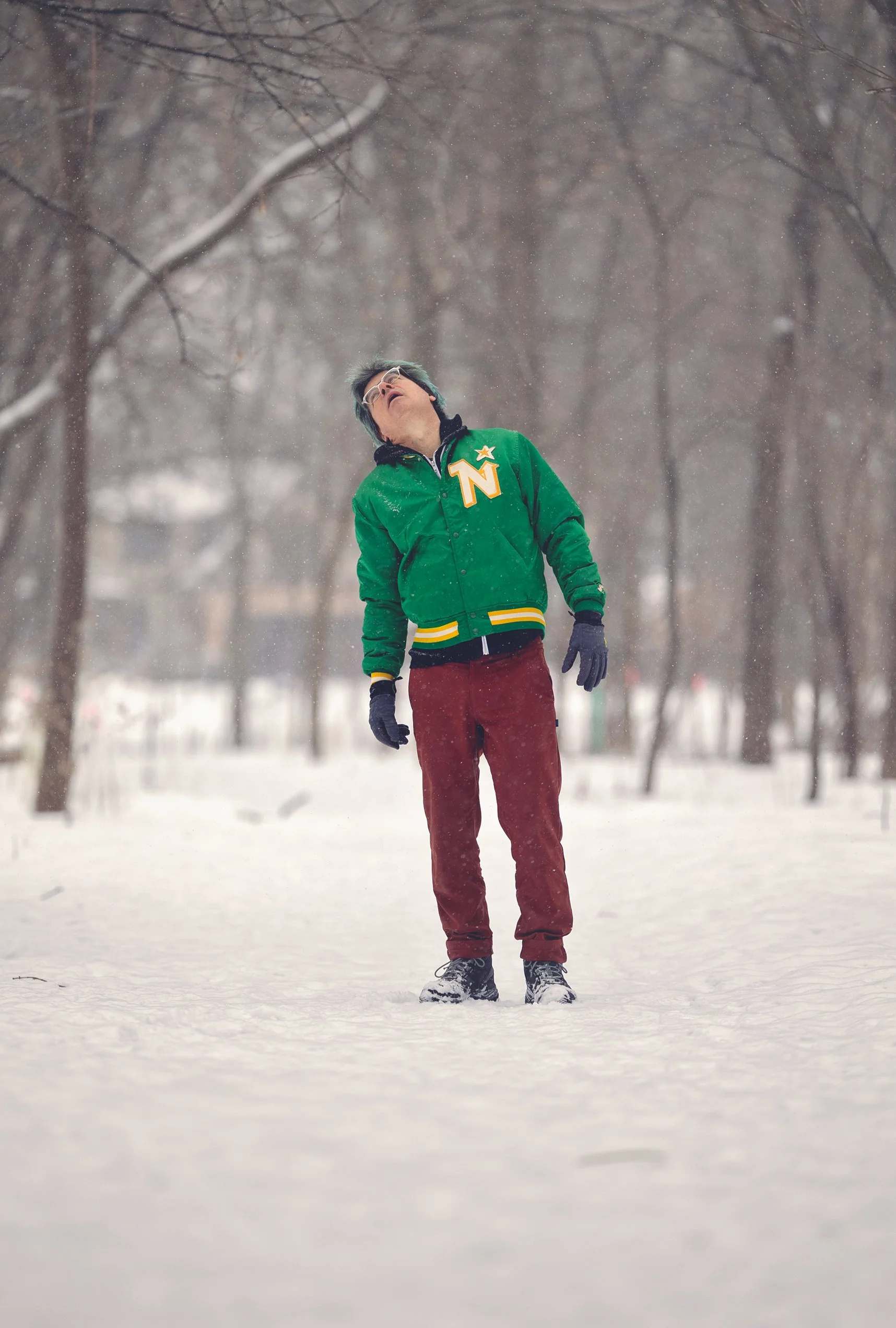 A person standing outdoors in a snow-covered park during snowfall, wearing a green jacket with a 'Northstars' patch, maroon pants, and black gloves, looking upward.