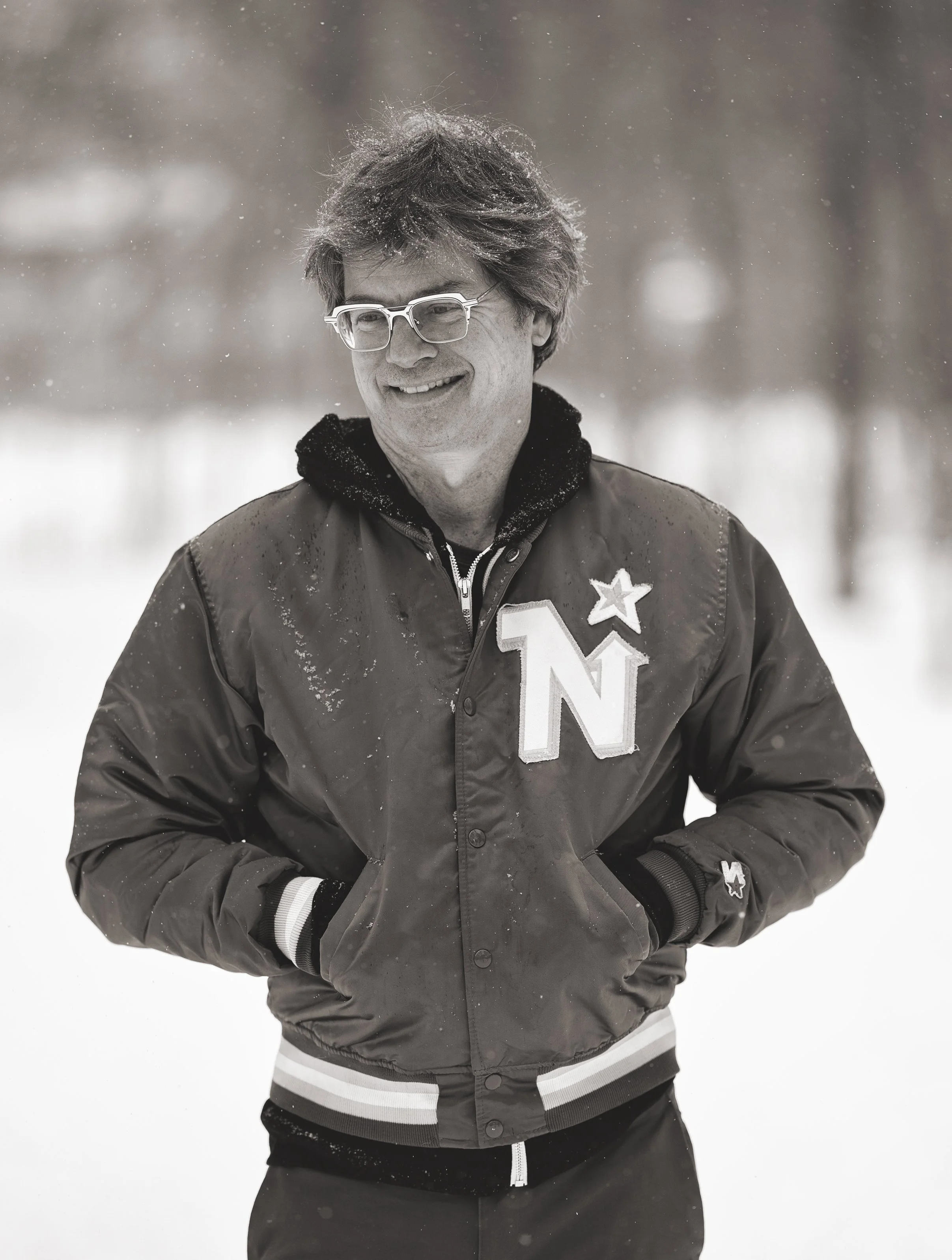 A man smiling outdoors in a snowy setting, wearing glasses, a varsity jacket with a large 'Northstars' patch, and hands in pockets.