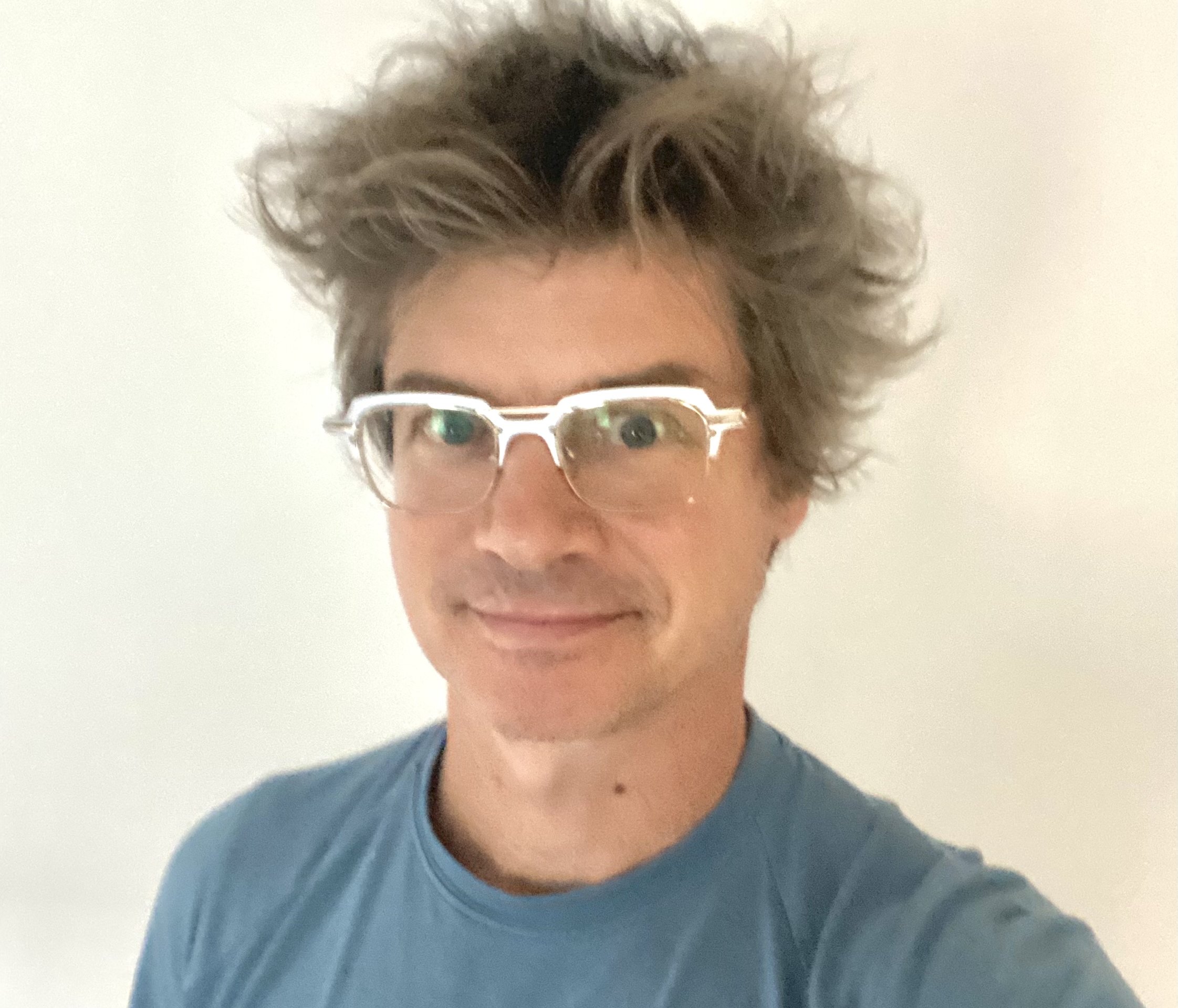 A man with light skin, messy brown hair, wearing clear glasses with a white frame, and a blue shirt, smiling against a plain light background.