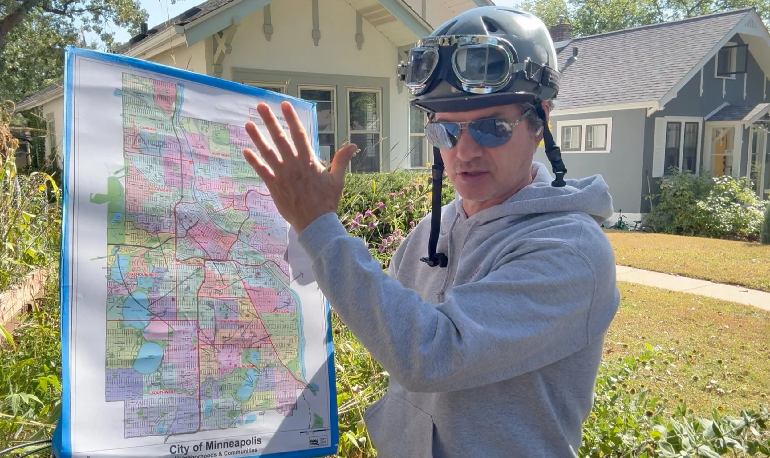 A man wearing a bicycle helmet and sunglasses, standing outdoors with houses and a garden in the background, pointing at a colorful map of Minneapolis city neighborhoods.