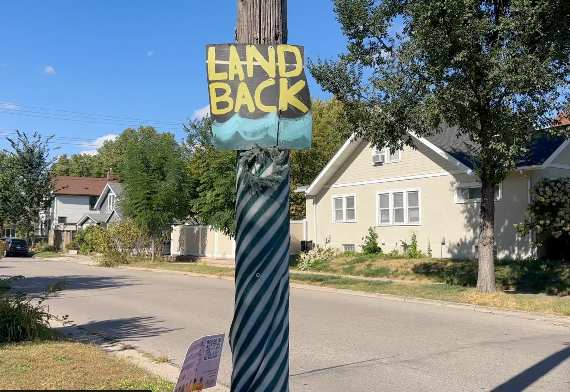 Hand-painted wooden sign on a pole that says 'LAND BACK' with a wave design at the bottom, on a residential street with houses, trees, and blue sky in the background.