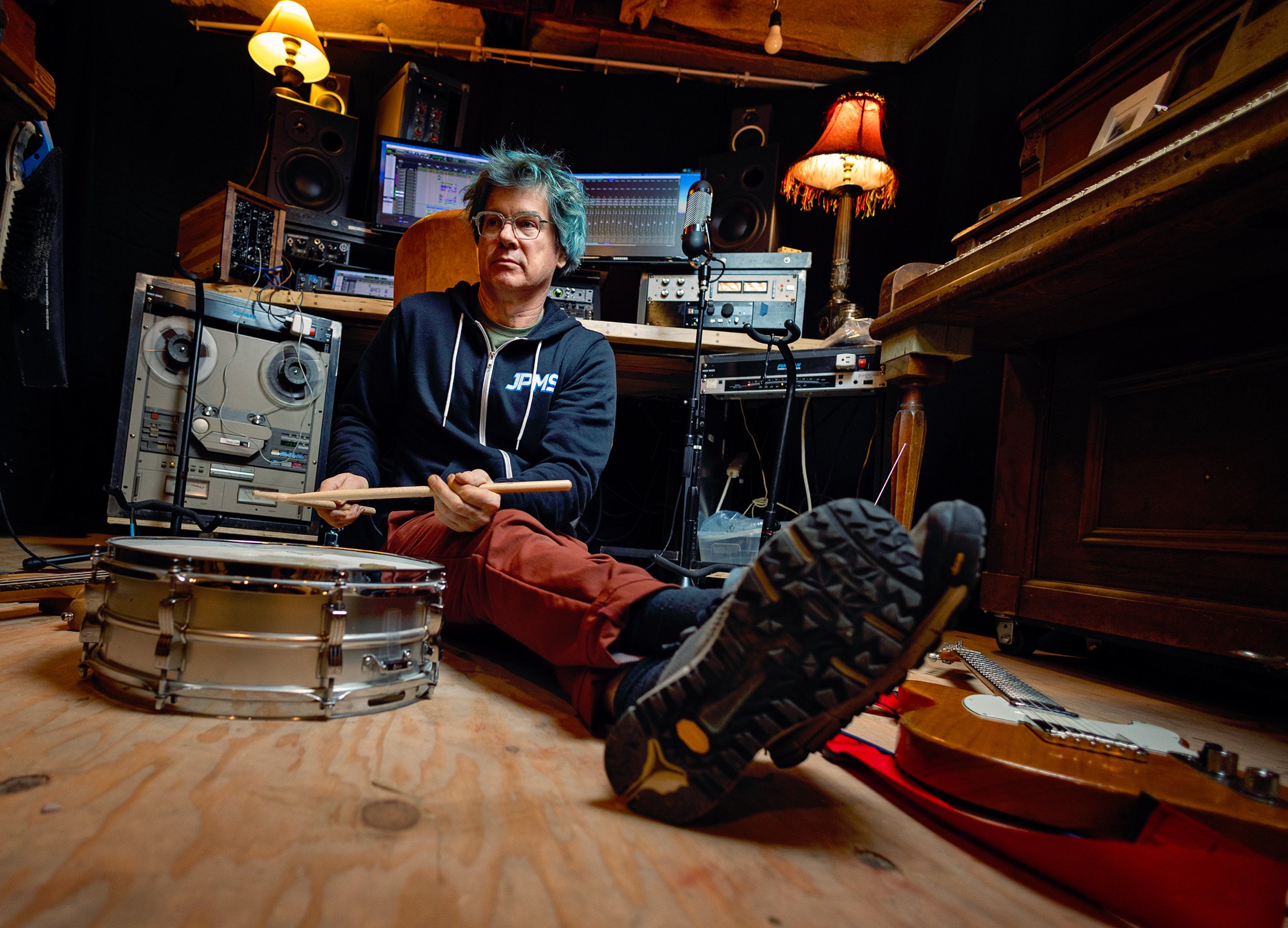 A man with curly hair and glasses sitting on the floor of a music studio, holding drumsticks, with a snare drum in front of him. The studio has various audio equipment, speakers, and computers behind him, with a warm lamp and a vintage-style lamp wit