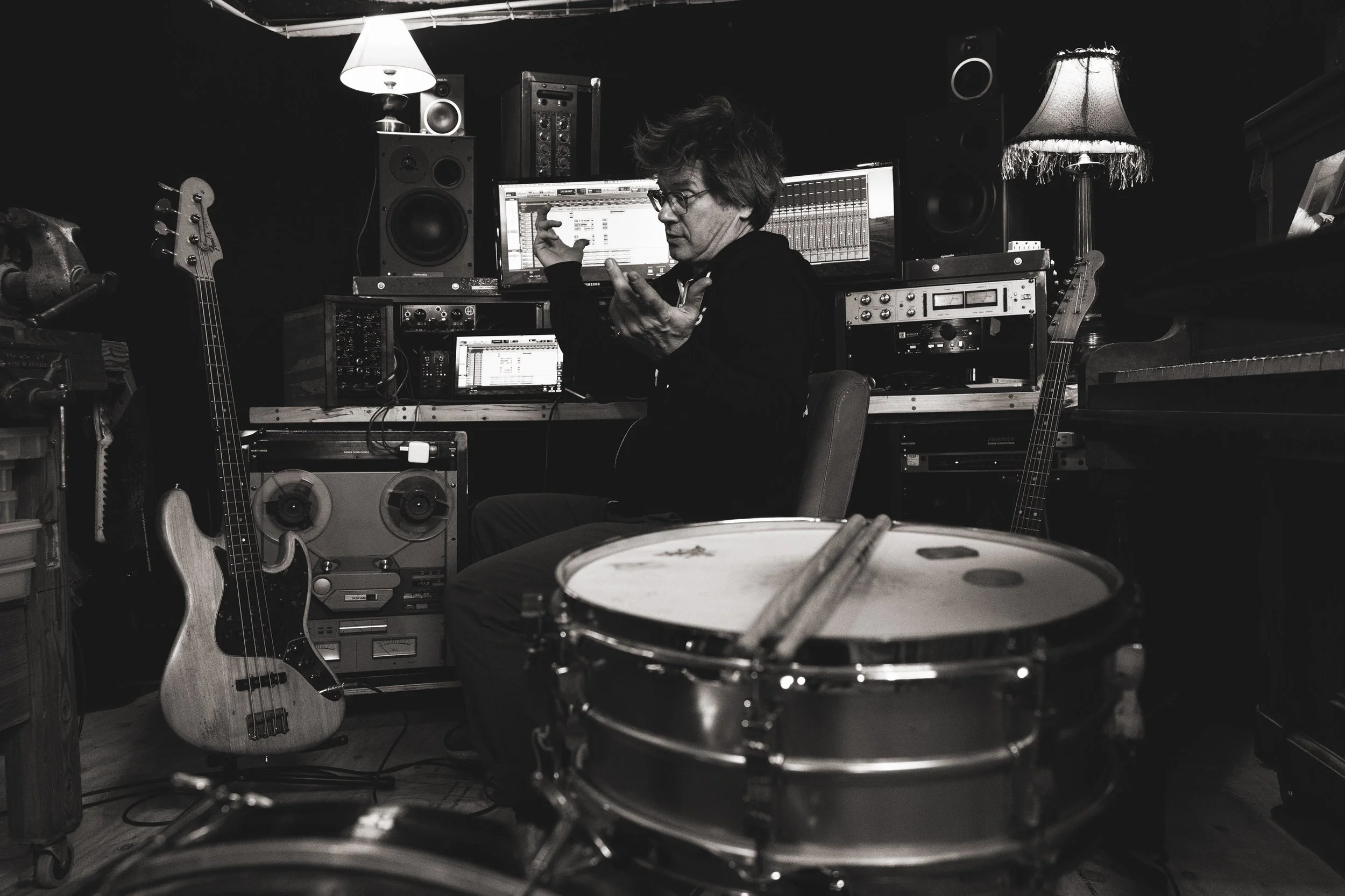 A man with glasses and messy hair in a music studio surrounded by musical equipment, including a bass guitar, drums, and studio monitors, working on music production in front of computer screens.